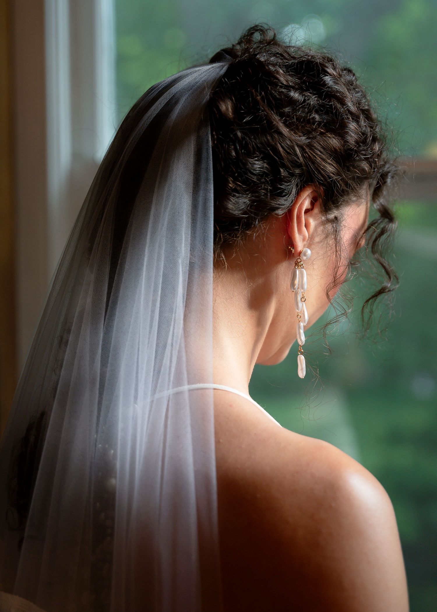Bride wearing a white veil and elegant drop earrings, with curly updo hairstyle, gazing out a window on her wedding day.