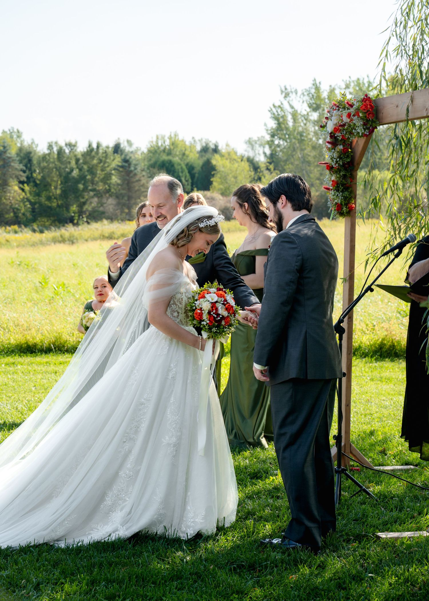 Outdoor wedding ceremony under a rustic wooden arch decorated with red flowers and willow branches.