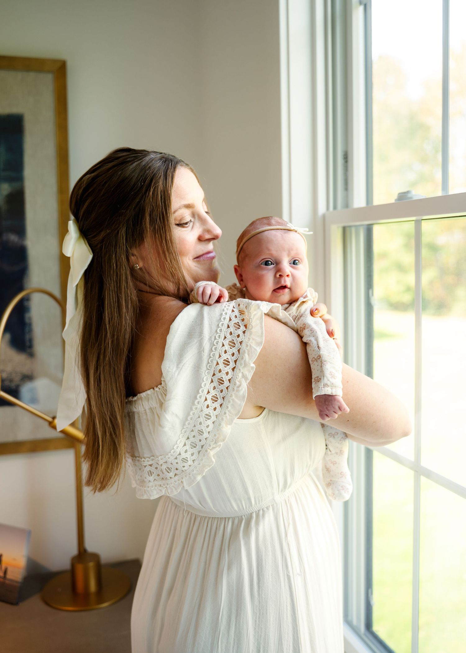 A woman in a white lace dress holds a baby near bright windows in a nursery setting.