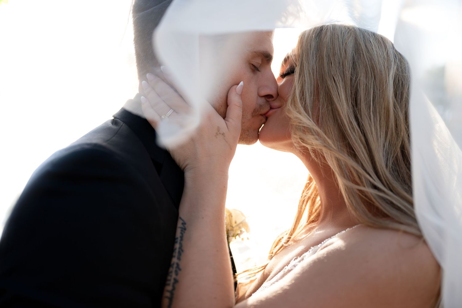 A bride and groom share a romantic kiss under a flowing white veil with soft golden backlight on their wedding day.