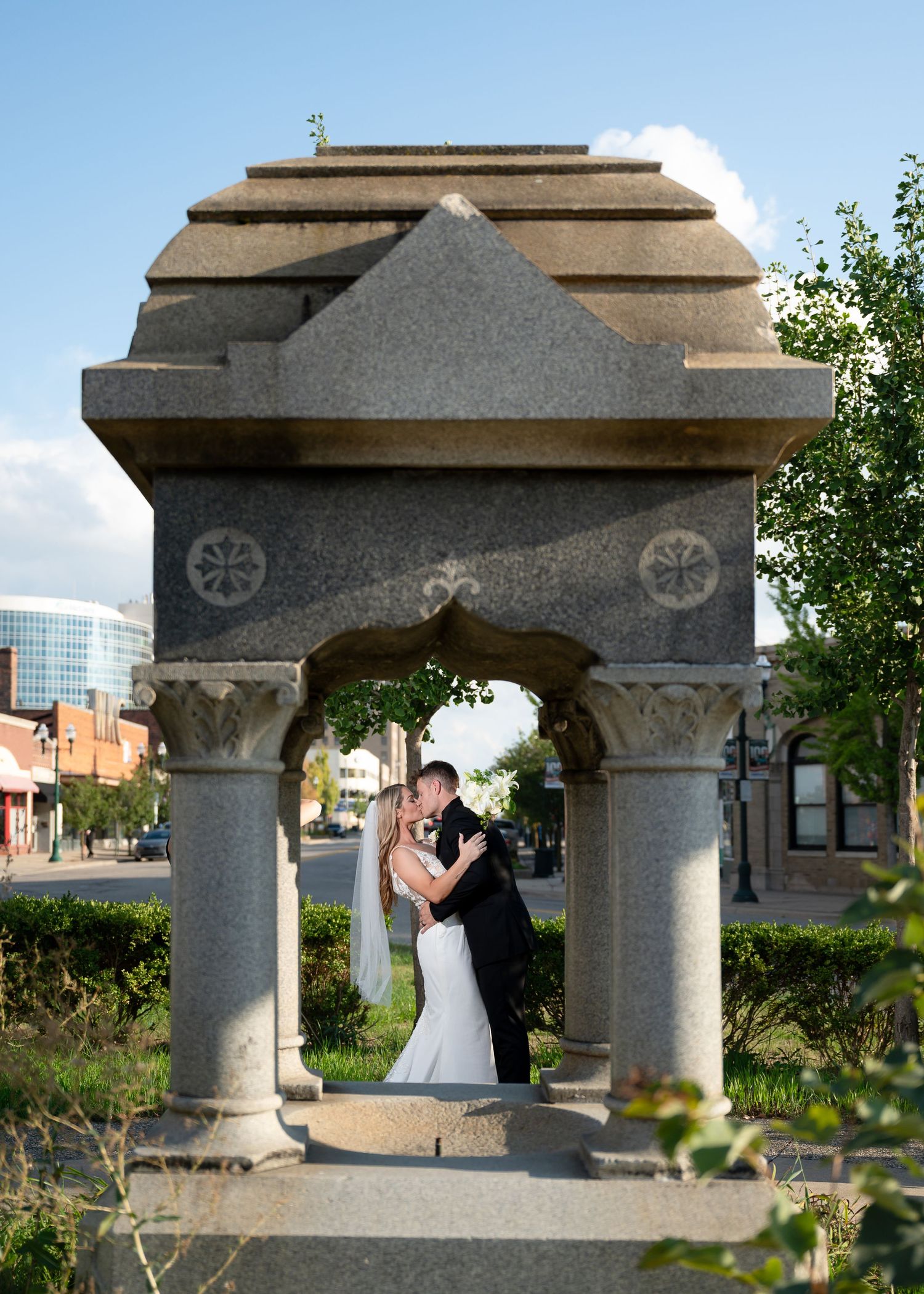 Bride and groom kissing under an ornate stone gazebo in an urban park setting on their wedding day.