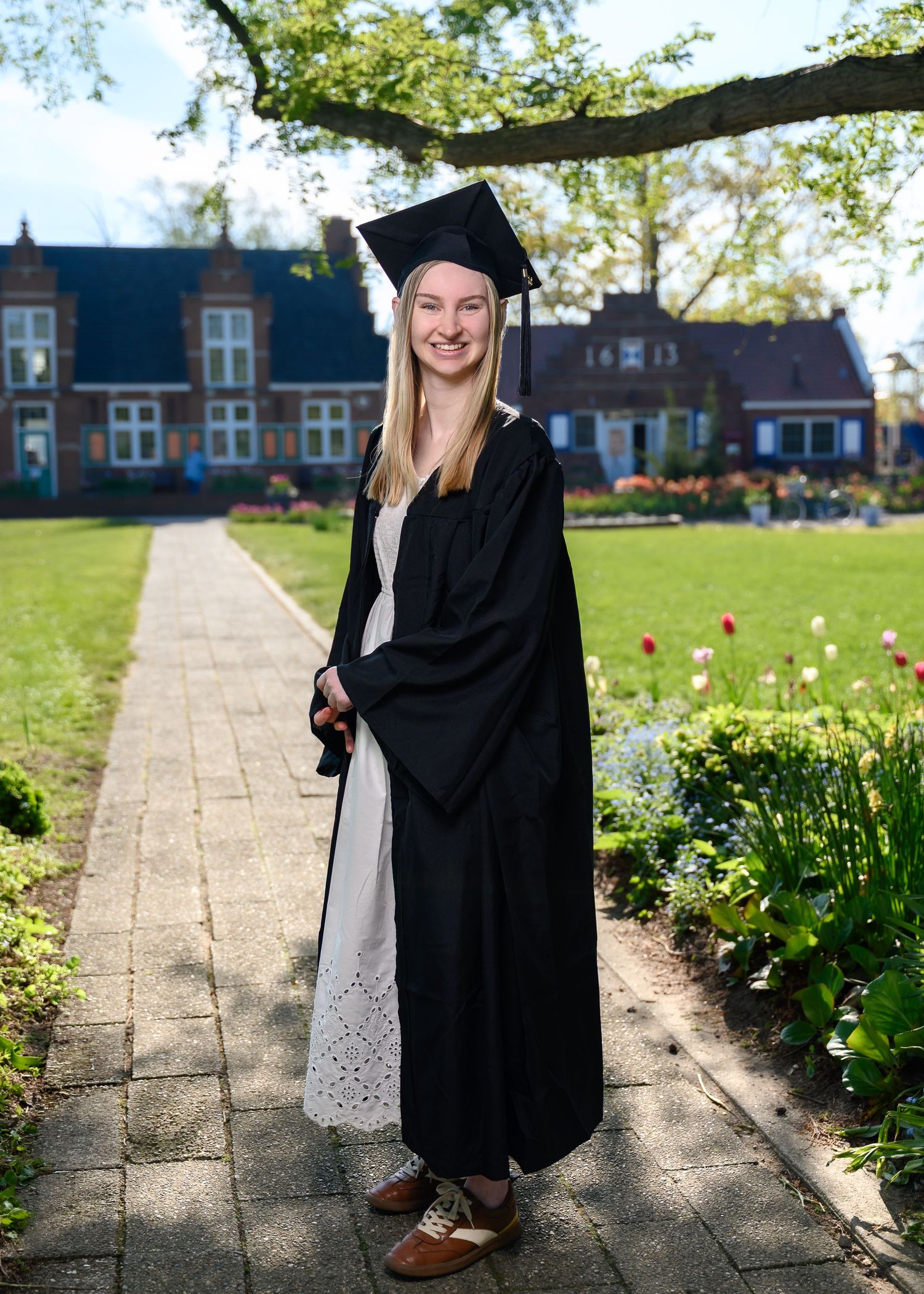 Smiling graduate in black cap and gown stands on garden path with tulips and brick building backdrop.