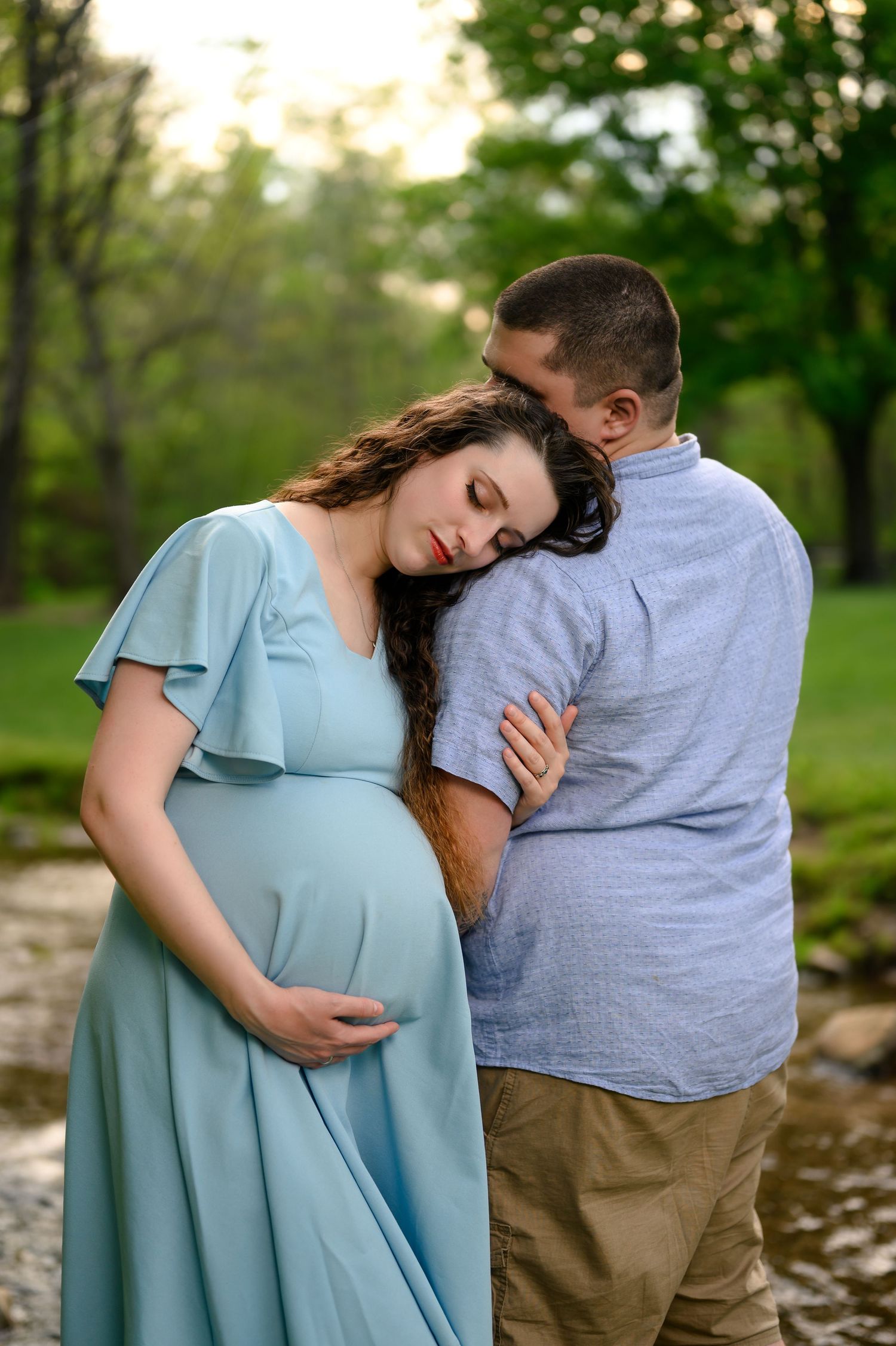 Pregnant woman in light blue dress leans on partner outdoors near stream, maternity photo session in green park setting.