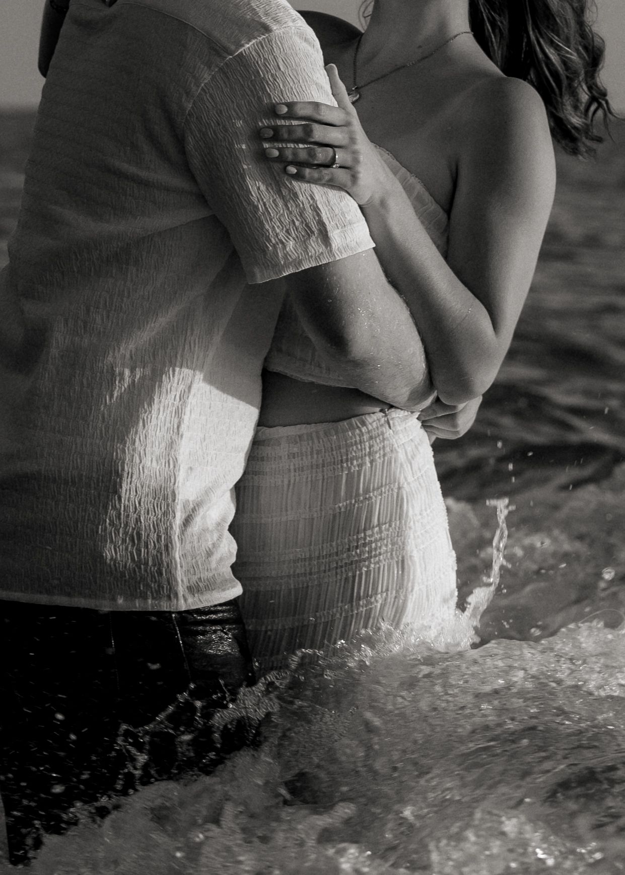 Black and white image of couple standing in ocean waves with water splashing around them.