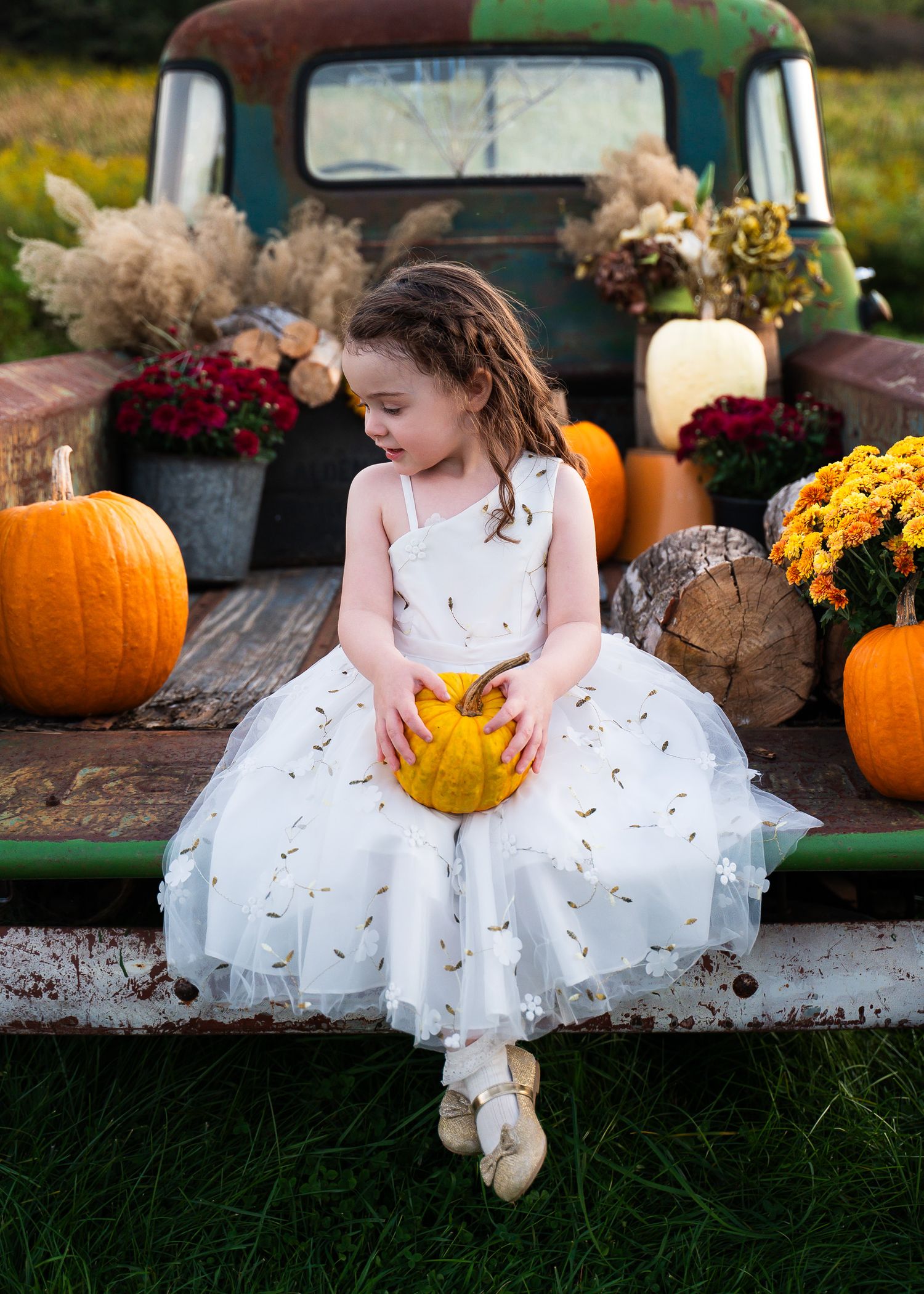 Young flower girl in white dress sits among autumn pumpkins and mums by vintage truck.