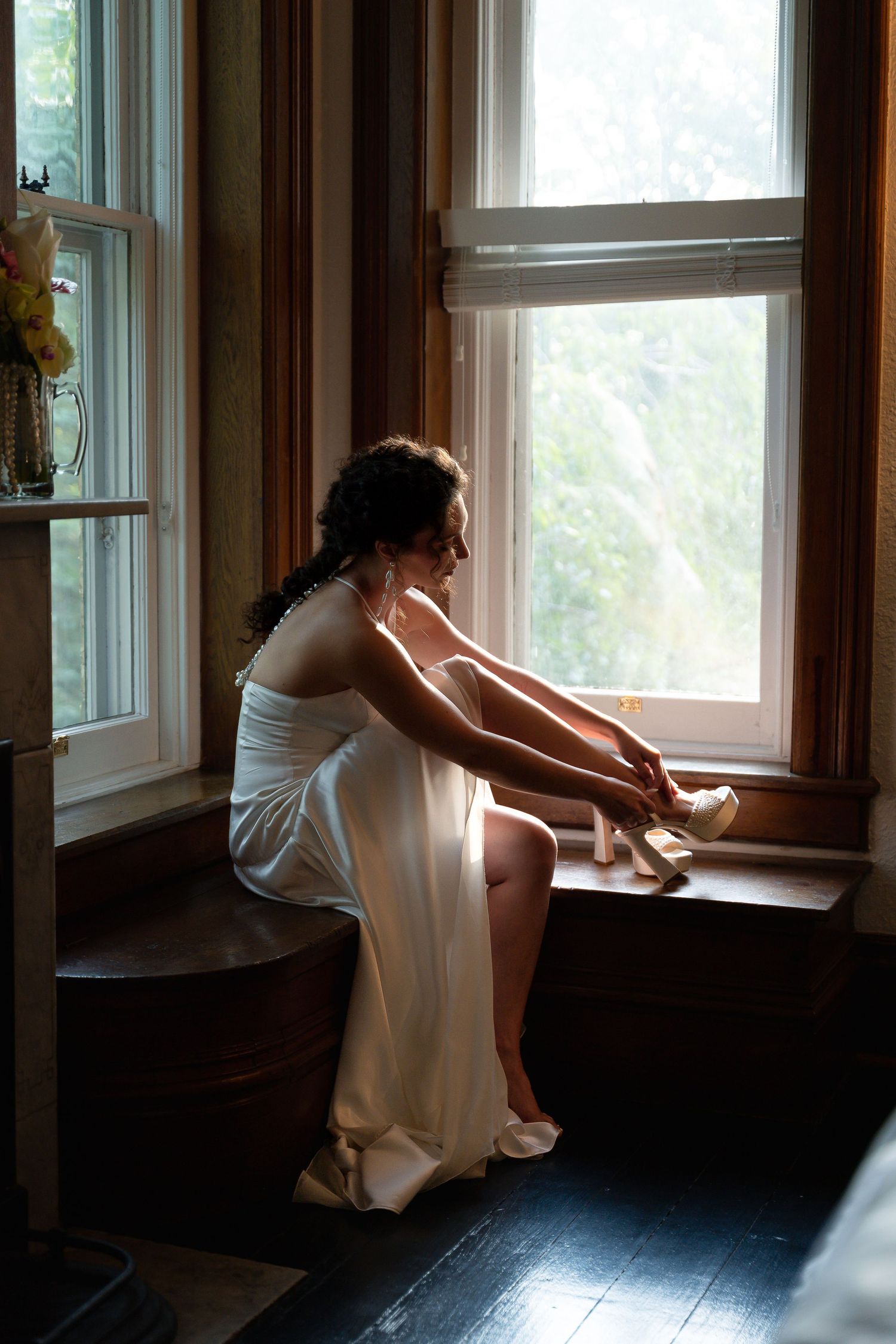 Bride in white satin gown sitting by window putting on heels, soft natural light, elegant wedding preparation moment.