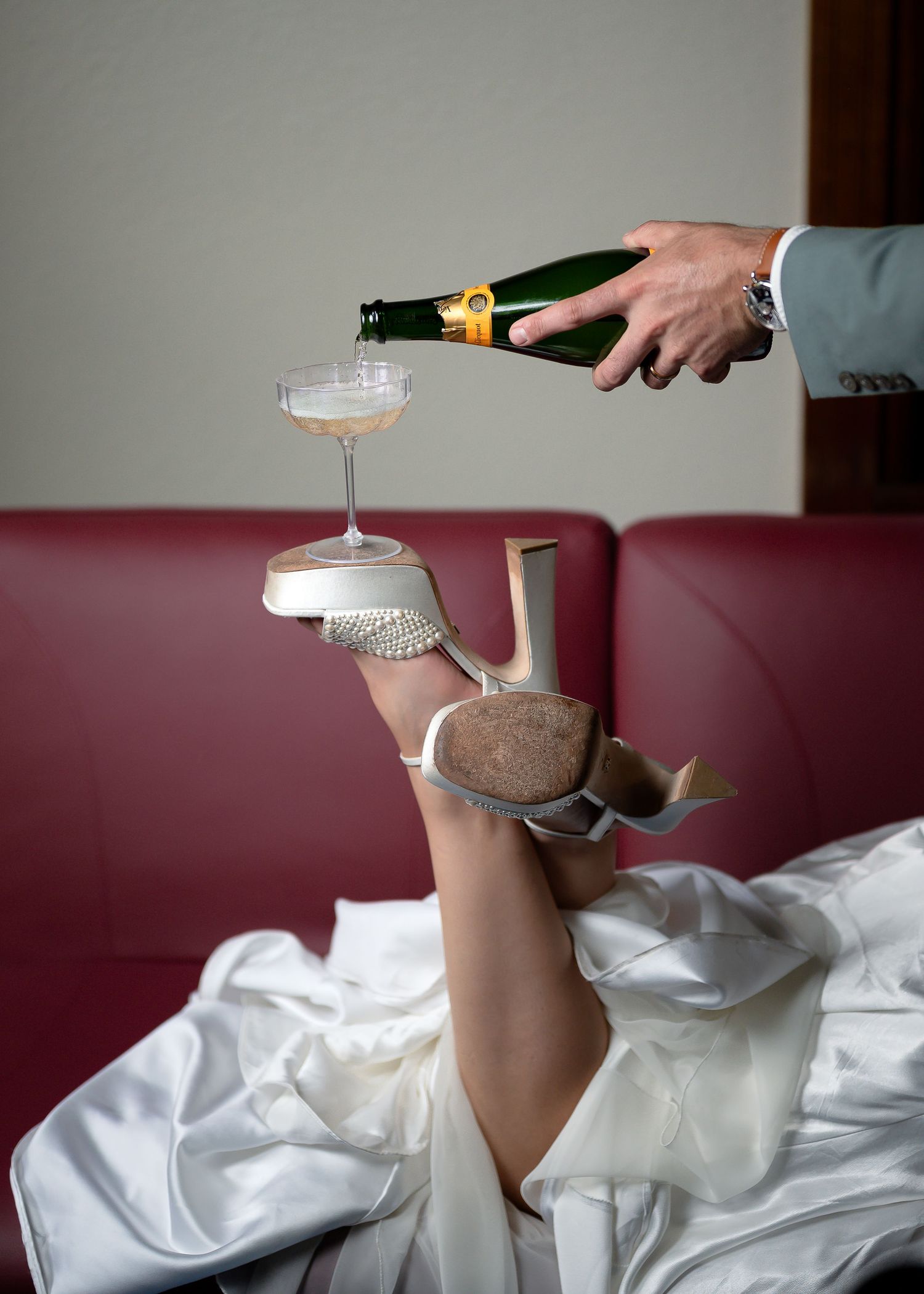 Person pouring champagne into a coupe glass balanced on a high heel shoe held up by a bride in a white dress.