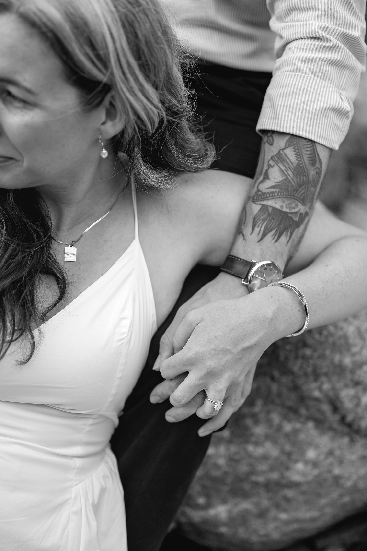 Black and white photo of a couple holding hands, woman in white dress with ring, man with tattoed arm and watch.