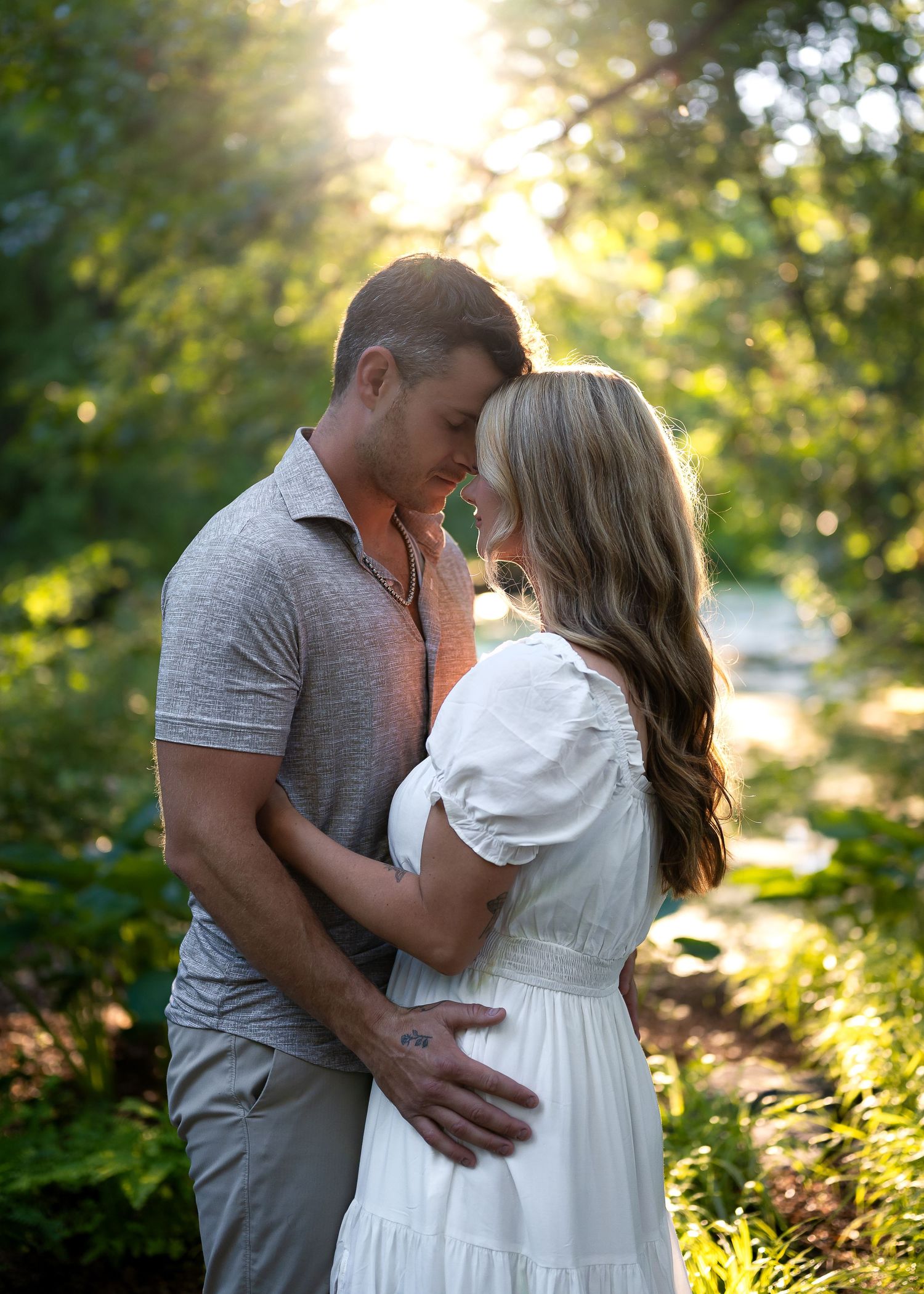 Couple sharing a romantic moment in a sunlit garden, man in gray shirt embracing woman in white dress.