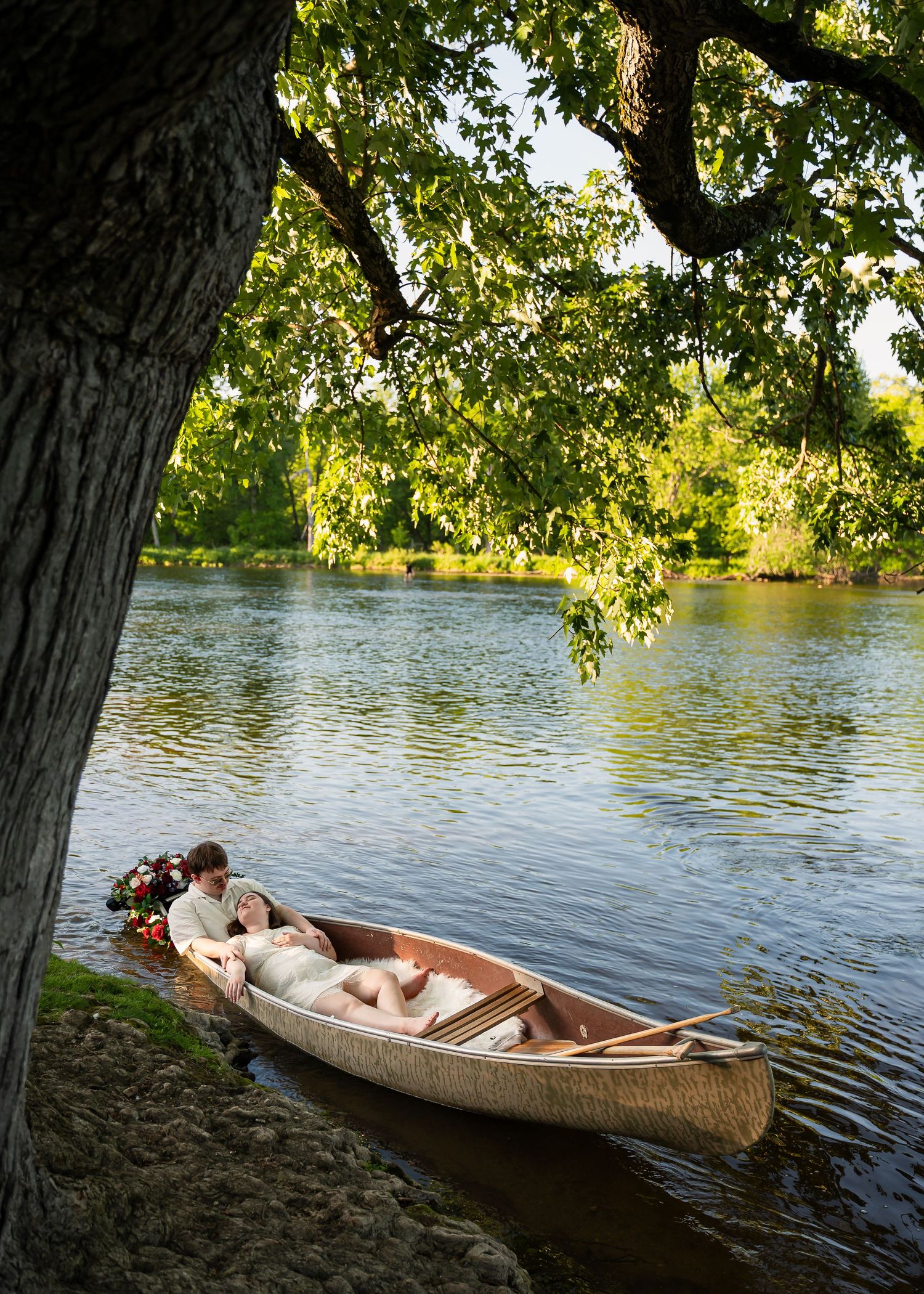 Romantic couple in wedding attire sharing a tender moment in a wooden canoe on a serene river under lush green trees.