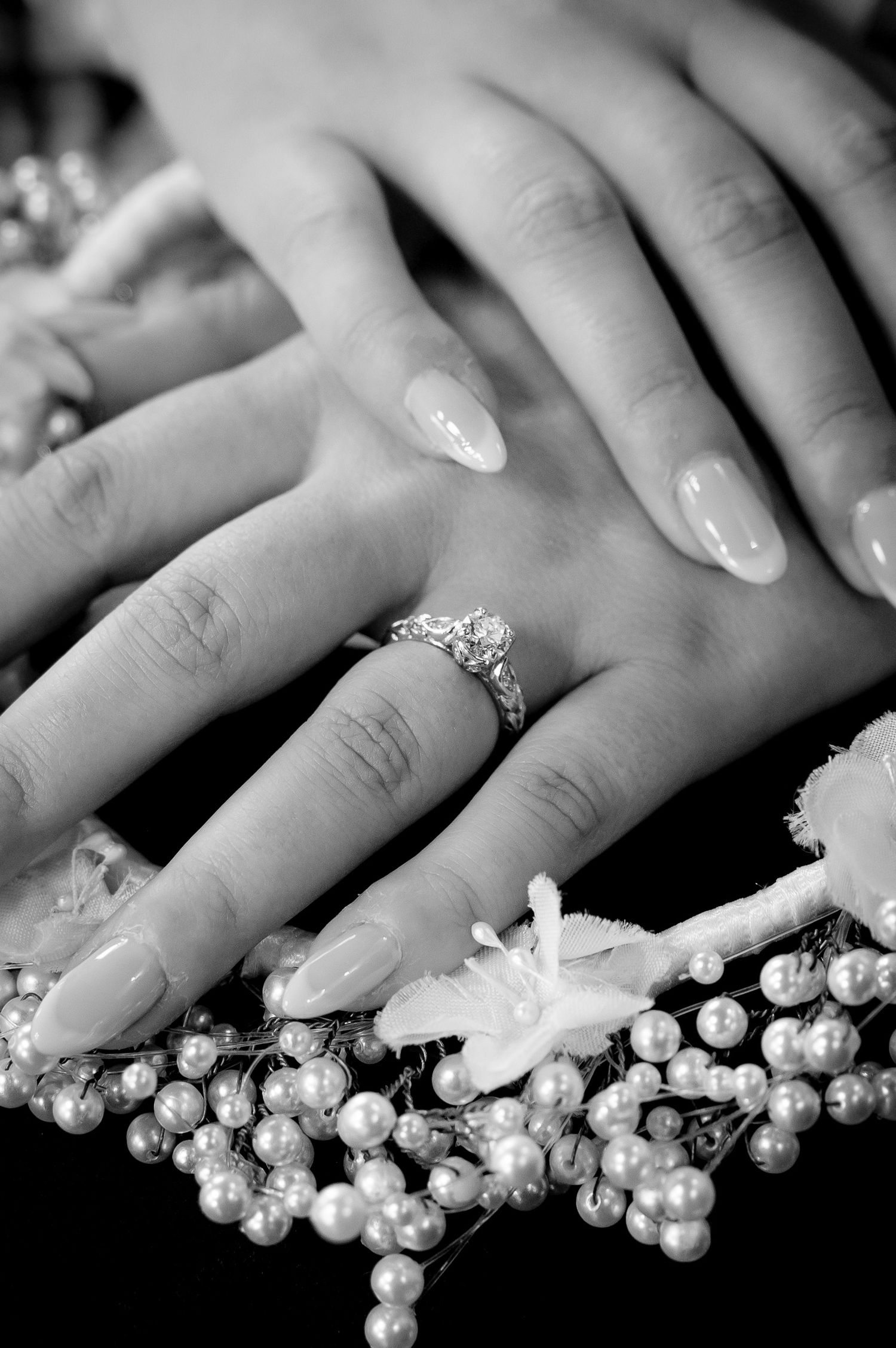 Black and white detail shot of manicured hands with engagement ring and pearl decorations.