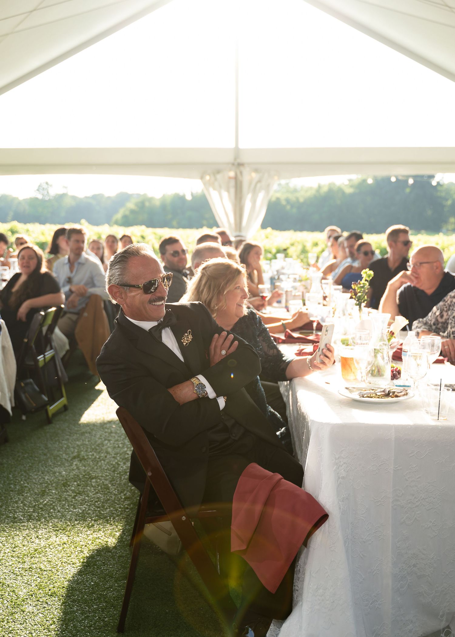 Guests seated at long white-draped tables under a tent at an outdoor wedding reception on a sunny day.