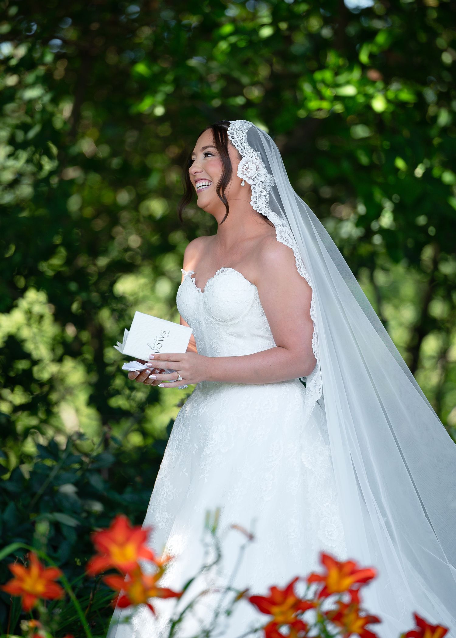 Smiling bride in white strapless gown and lace veil holds a card outdoors among lush green trees and orange flowers.