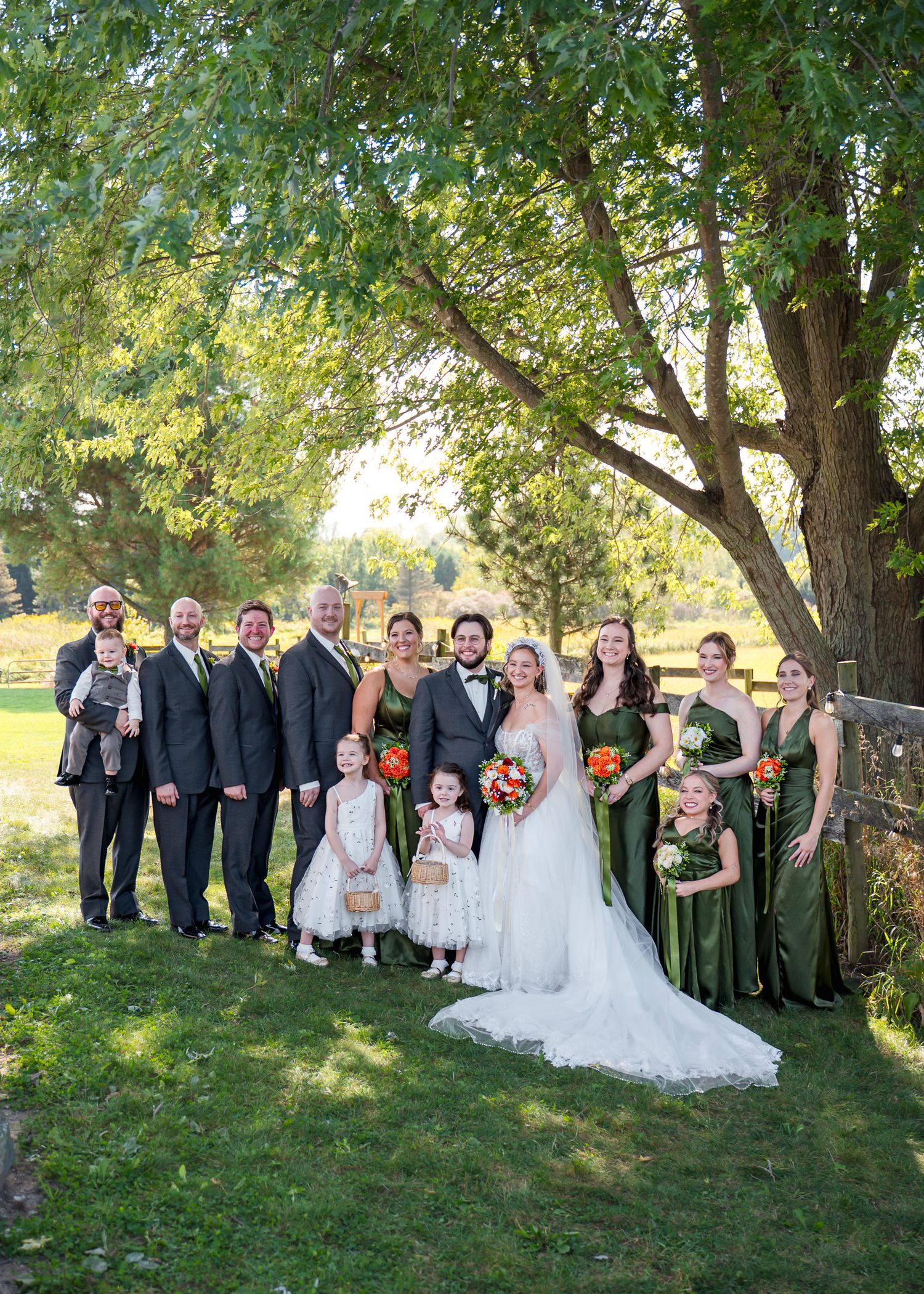 Large wedding party poses under green trees for formal group photo.