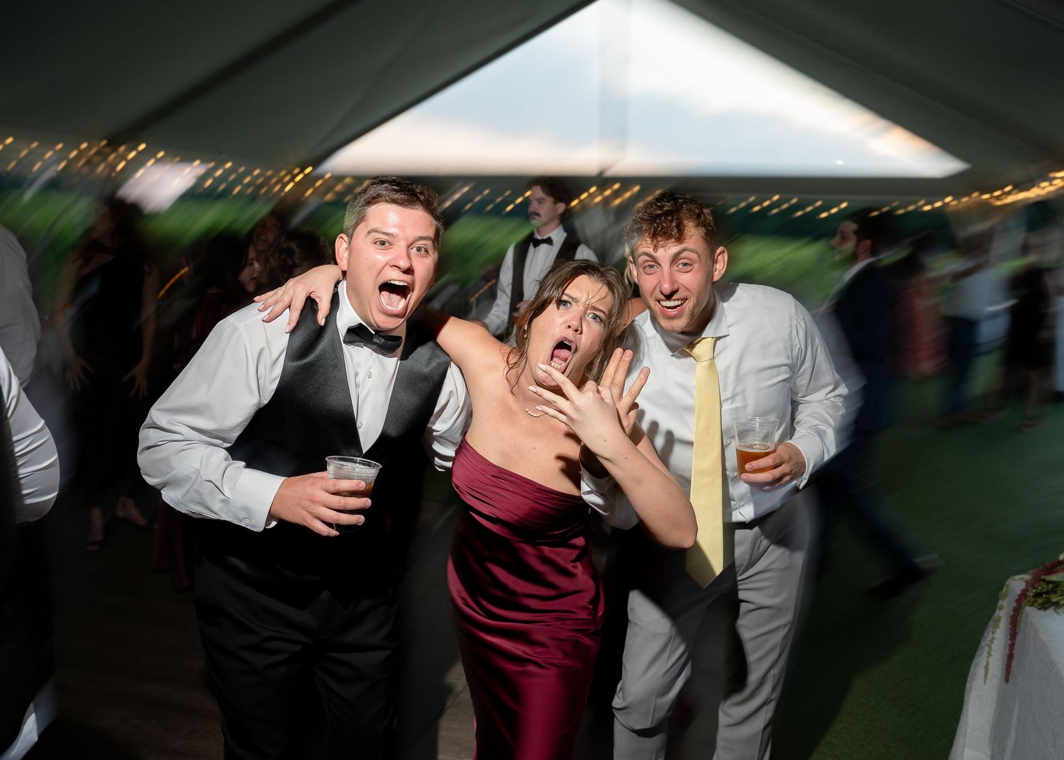 Three friends pose dramatically at a wedding reception under a white tent, wearing formal attire.
