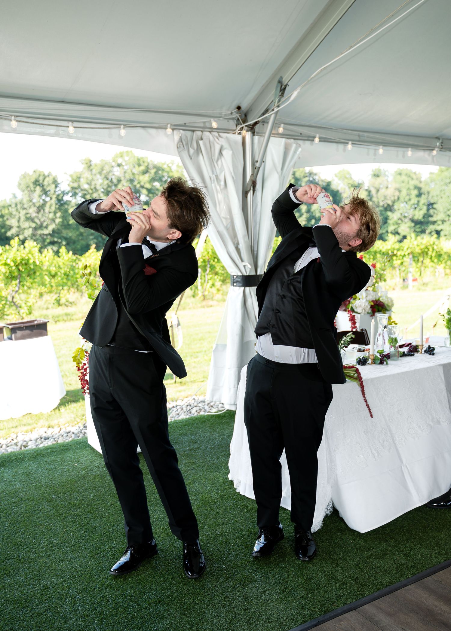 Two groomsmen in black tuxedos doing shots under a white wedding tent with vineyard views.