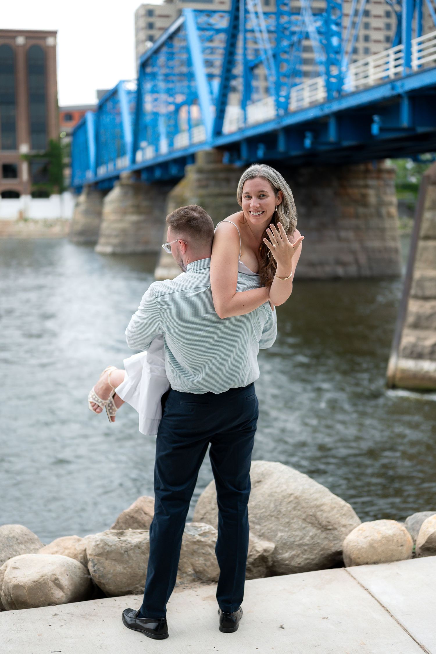 Man carrying woman piggyback near river with blue bridge, woman smiling and showing engagement ring.