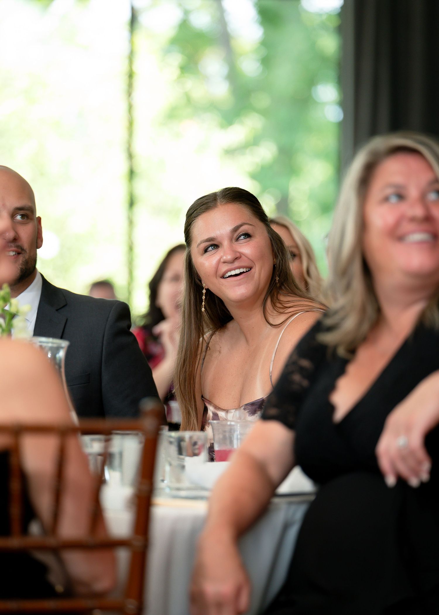 Smiling woman in white dress enjoys a wedding reception dinner with guests seated at decorated tables.