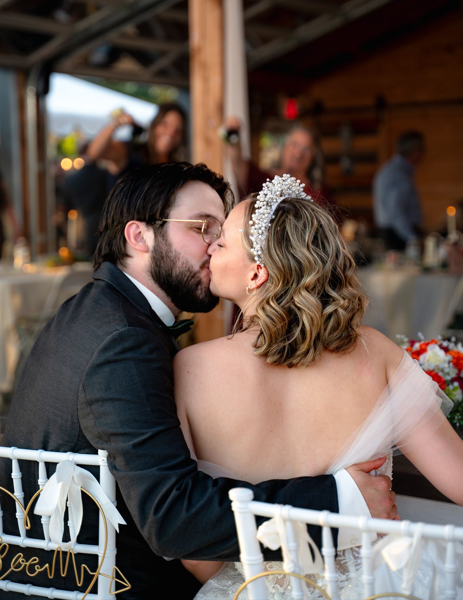 A wedding couple shares an intimate kiss at their reception while seated in white chairs.