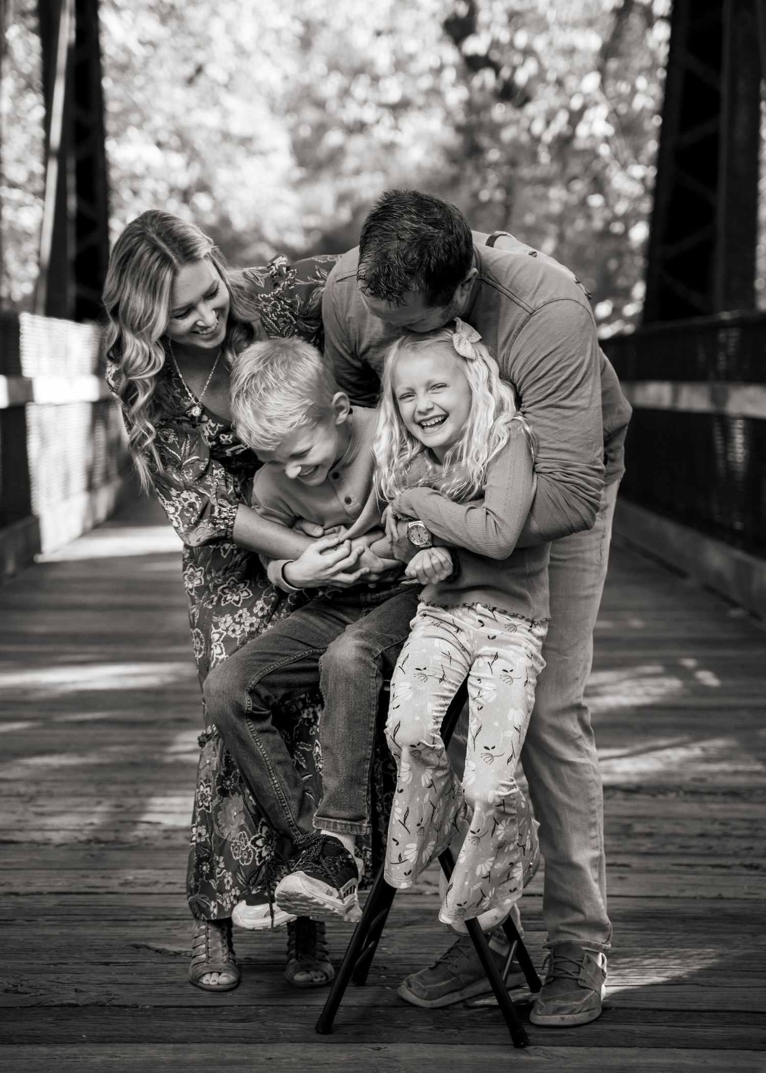 Black and white photo of family sharing playful moment on wooden bridge.