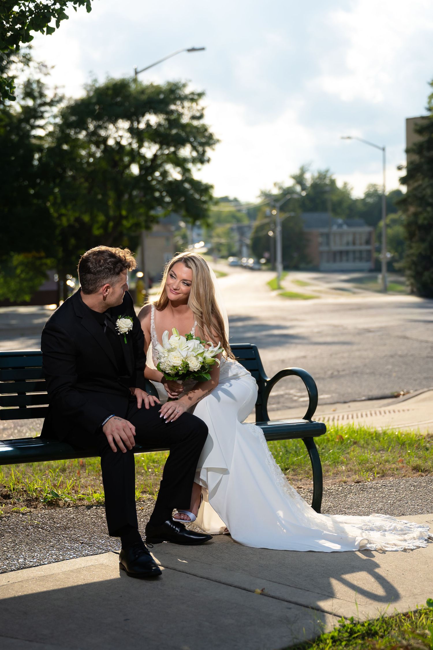 Bride and groom sitting on a park bench in wedding attire, bride holding white bouquet, sharing an intimate moment outdoors.