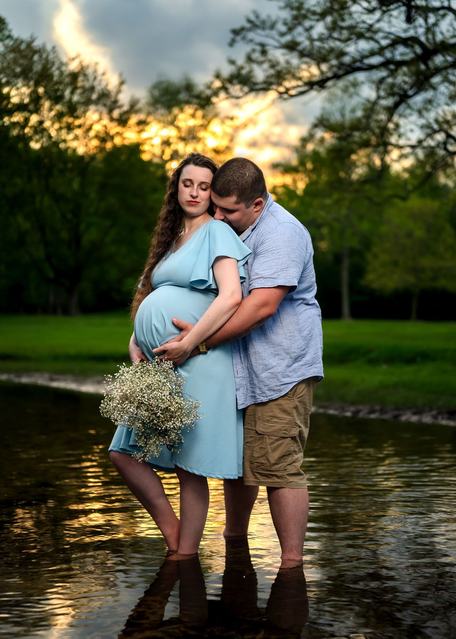 Pregnant couple standing in water at sunset, woman in blue dress holding flowers, man embracing her baby bump.