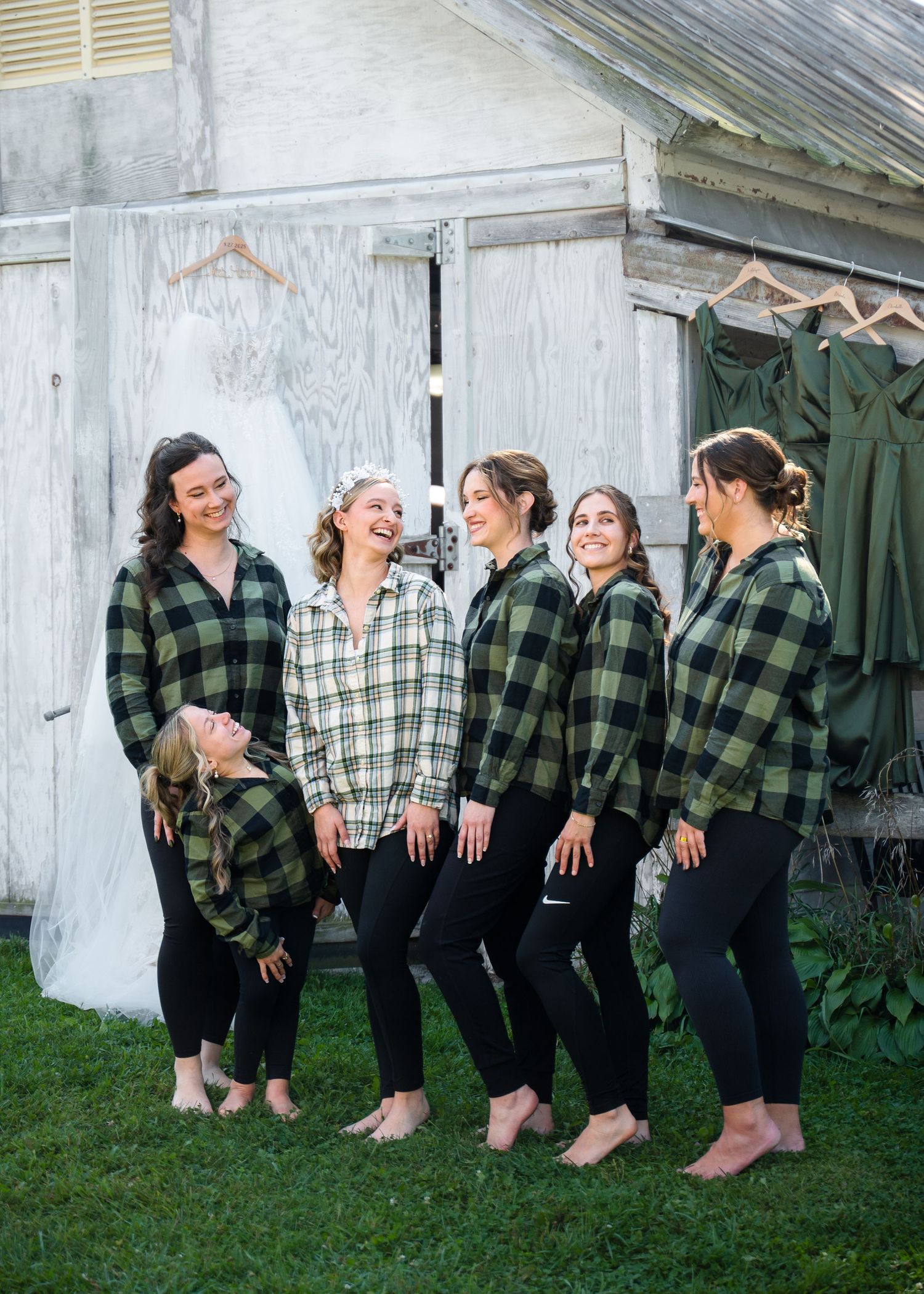 Wedding party in matching green plaid shirts poses and laughs together against a rustic white building.