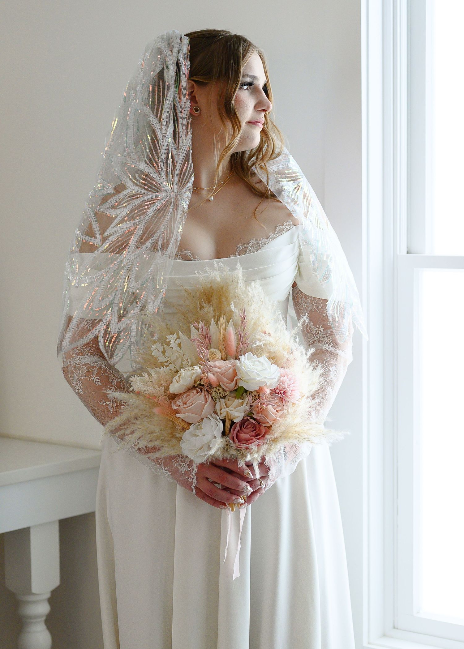Bride in white lace wedding dress and floral veil holding pink and white bouquet, standing by bright window.