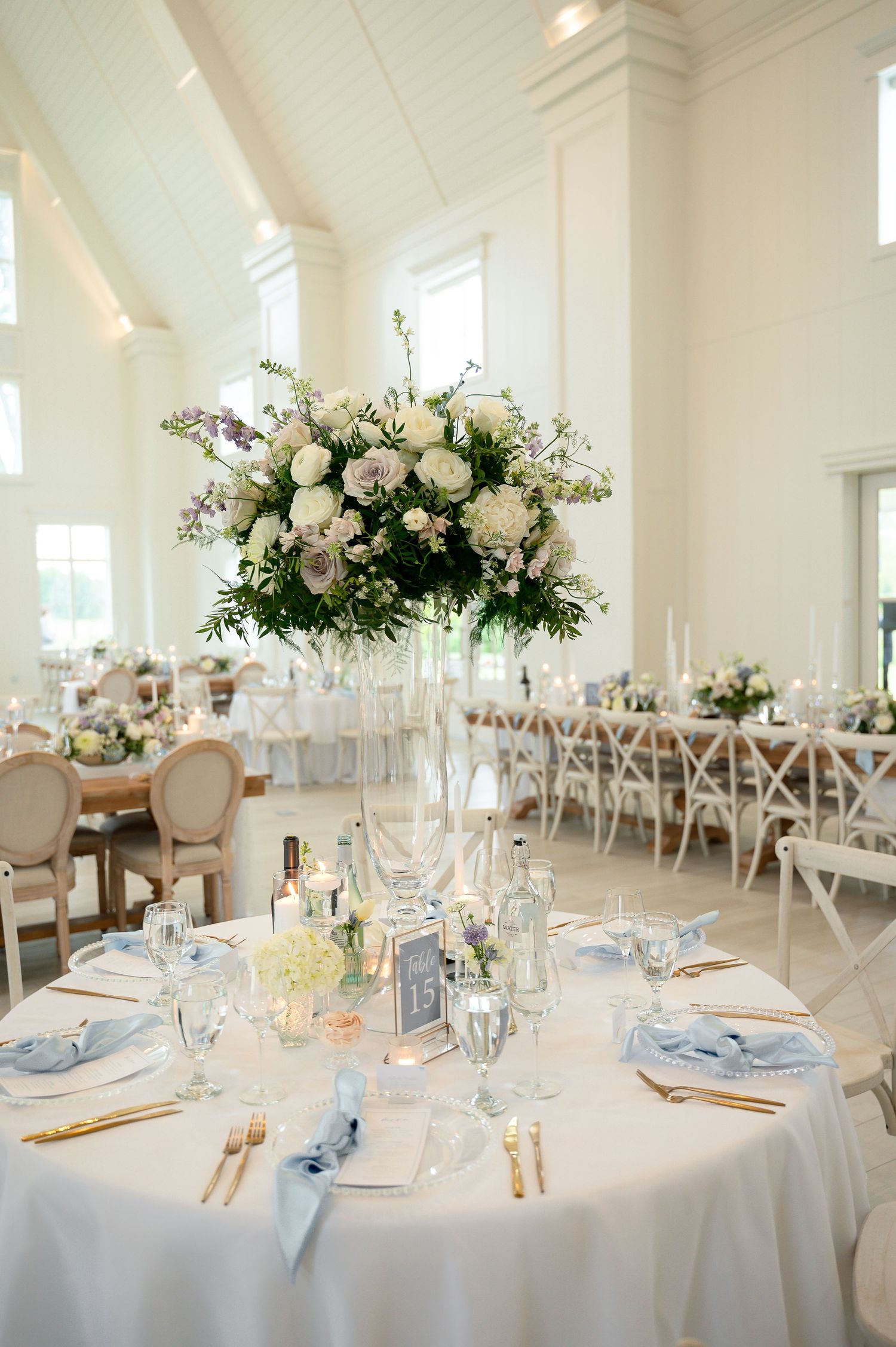 Elegant wedding reception with tall floral centerpiece of white and lavender blooms, blue napkins, and gold cutlery.