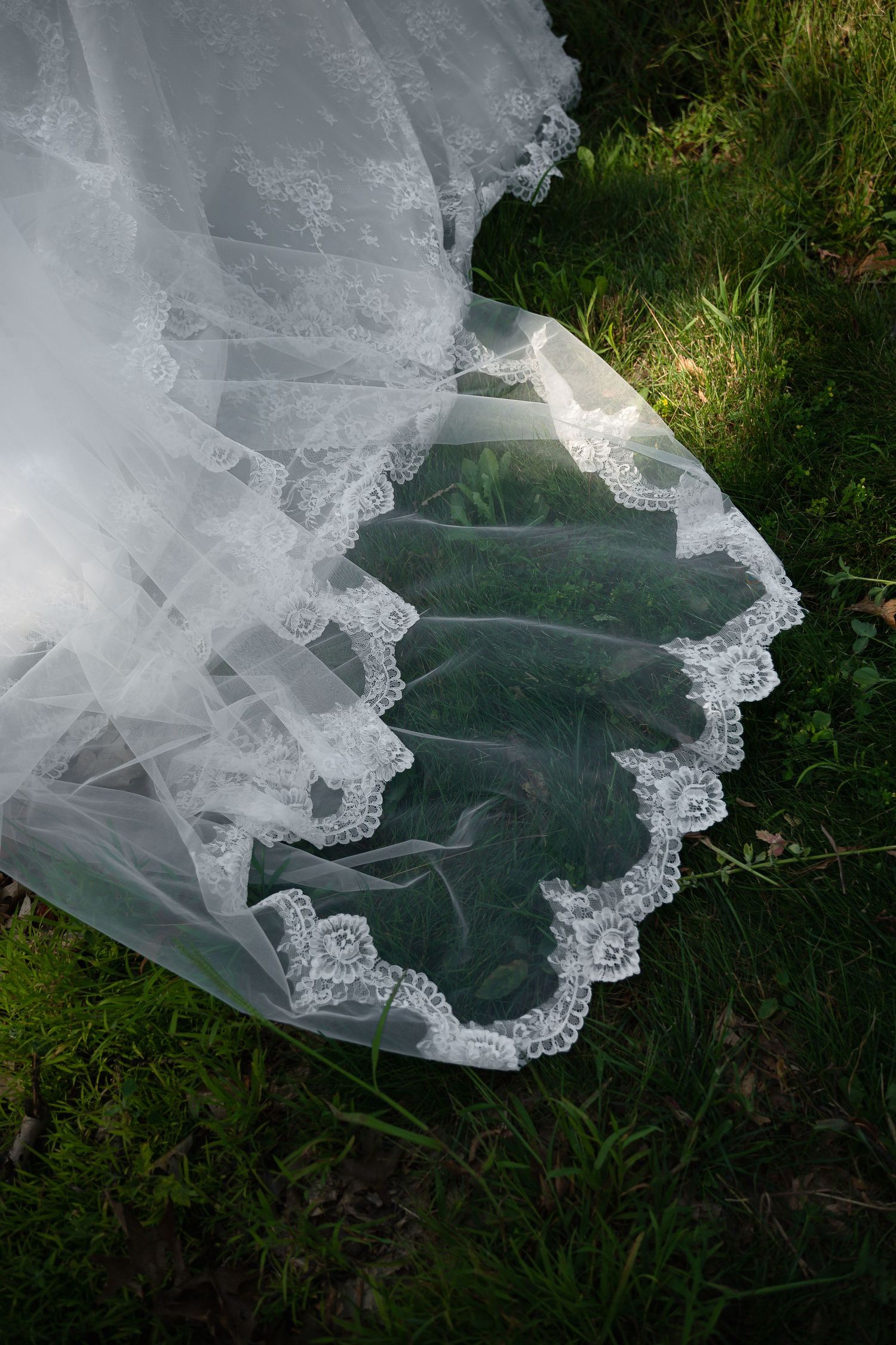 White lace wedding veil with floral trim draped elegantly over green grass outdoors.