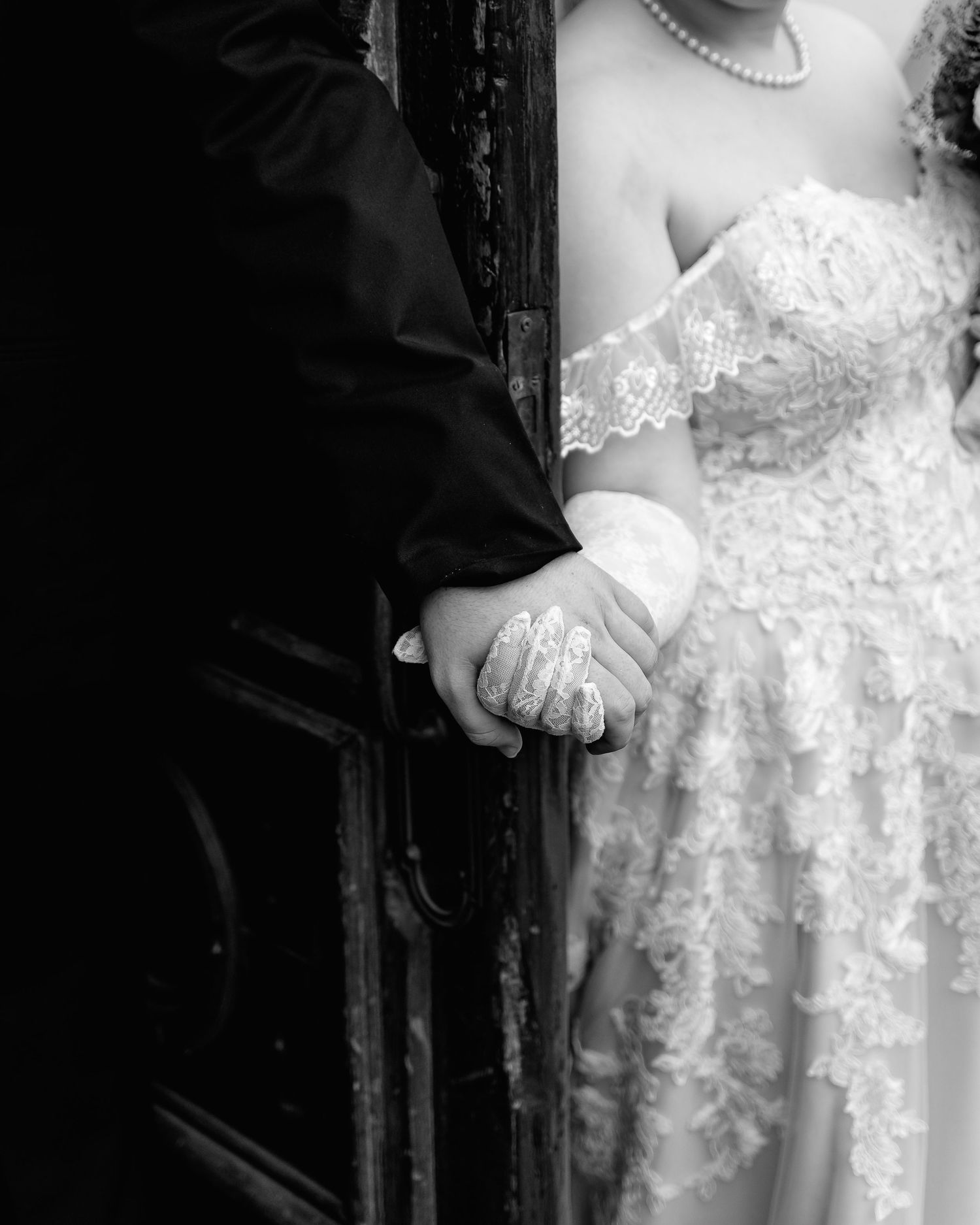 Black and white photo of a bride and groom holding hands, showcasing lace wedding dress and diamond ring.