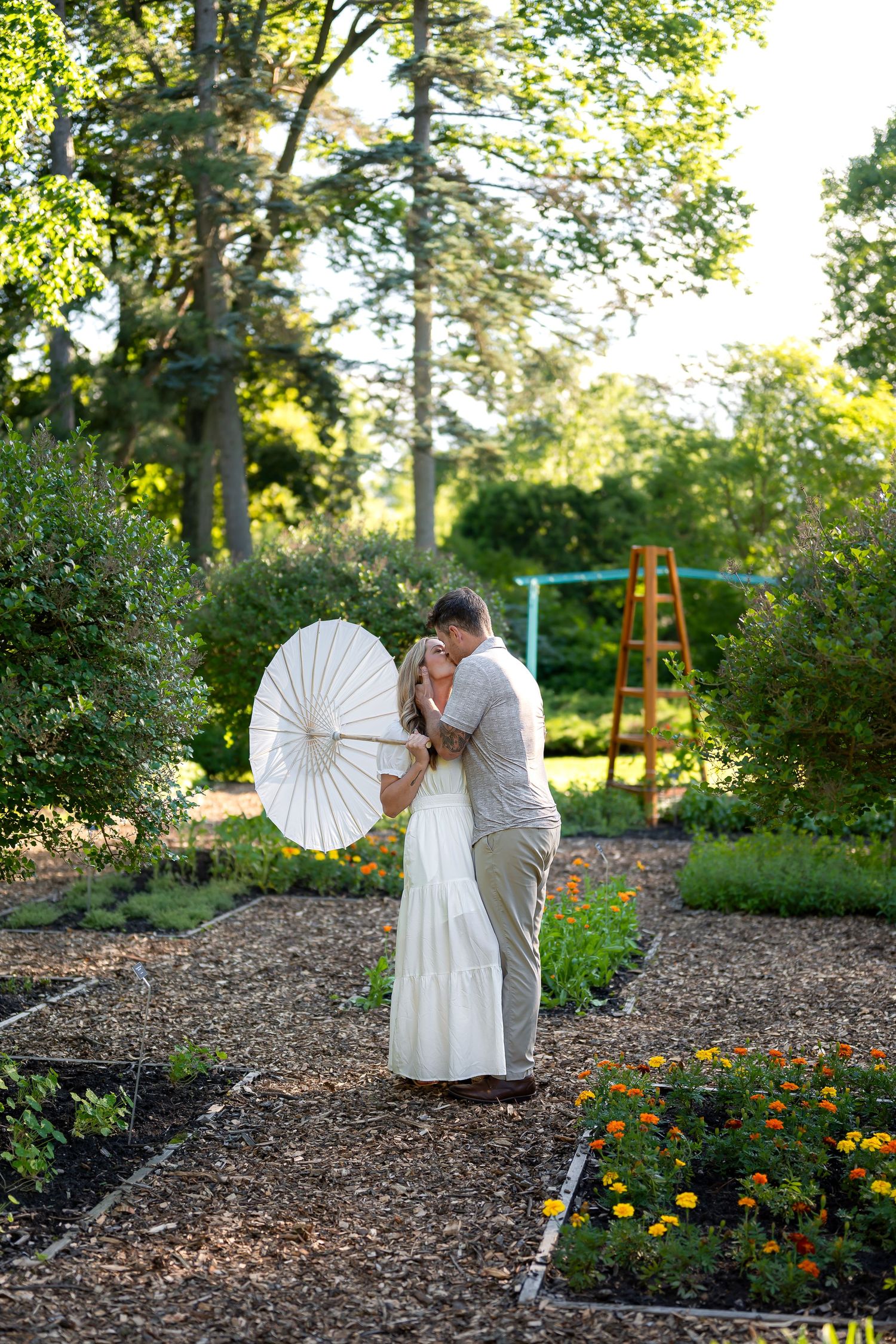 Couple sharing a romantic kiss in a lush garden, woman in white dress holding a parasol umbrella.