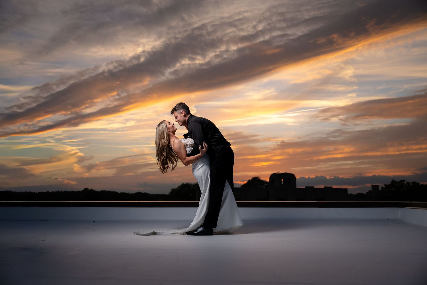 Couple sharing a romantic dip kiss at sunset on a rooftop, with dramatic golden clouds in the background.