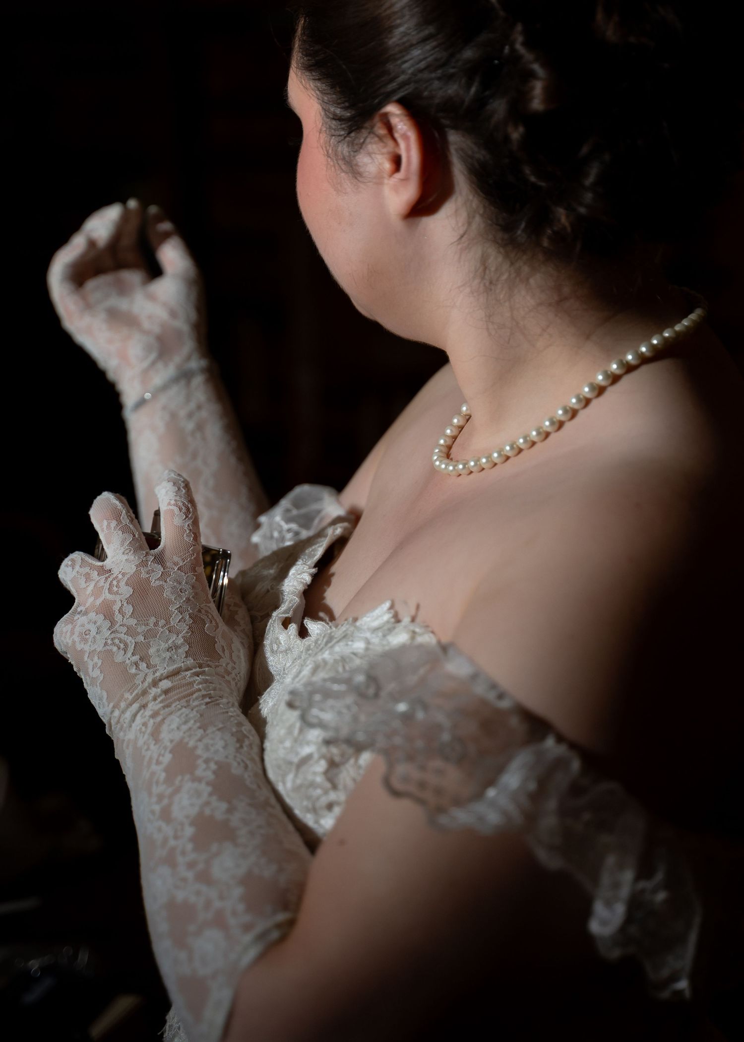 Bride in white lace gloves and pearl necklace adjusting her wedding dress in a dark, elegant setting.