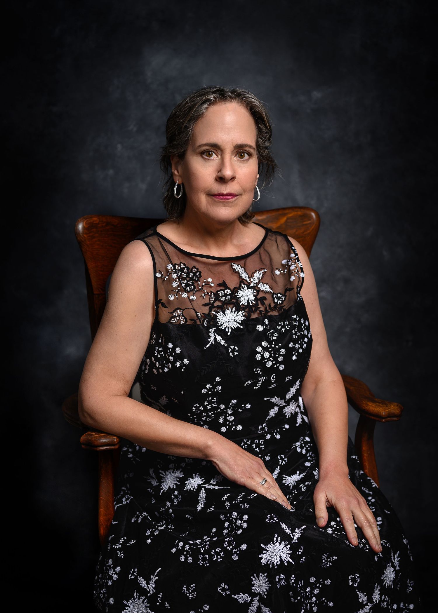 Elegant woman in black floral embroidered dress seated in wooden chair against dark studio background.