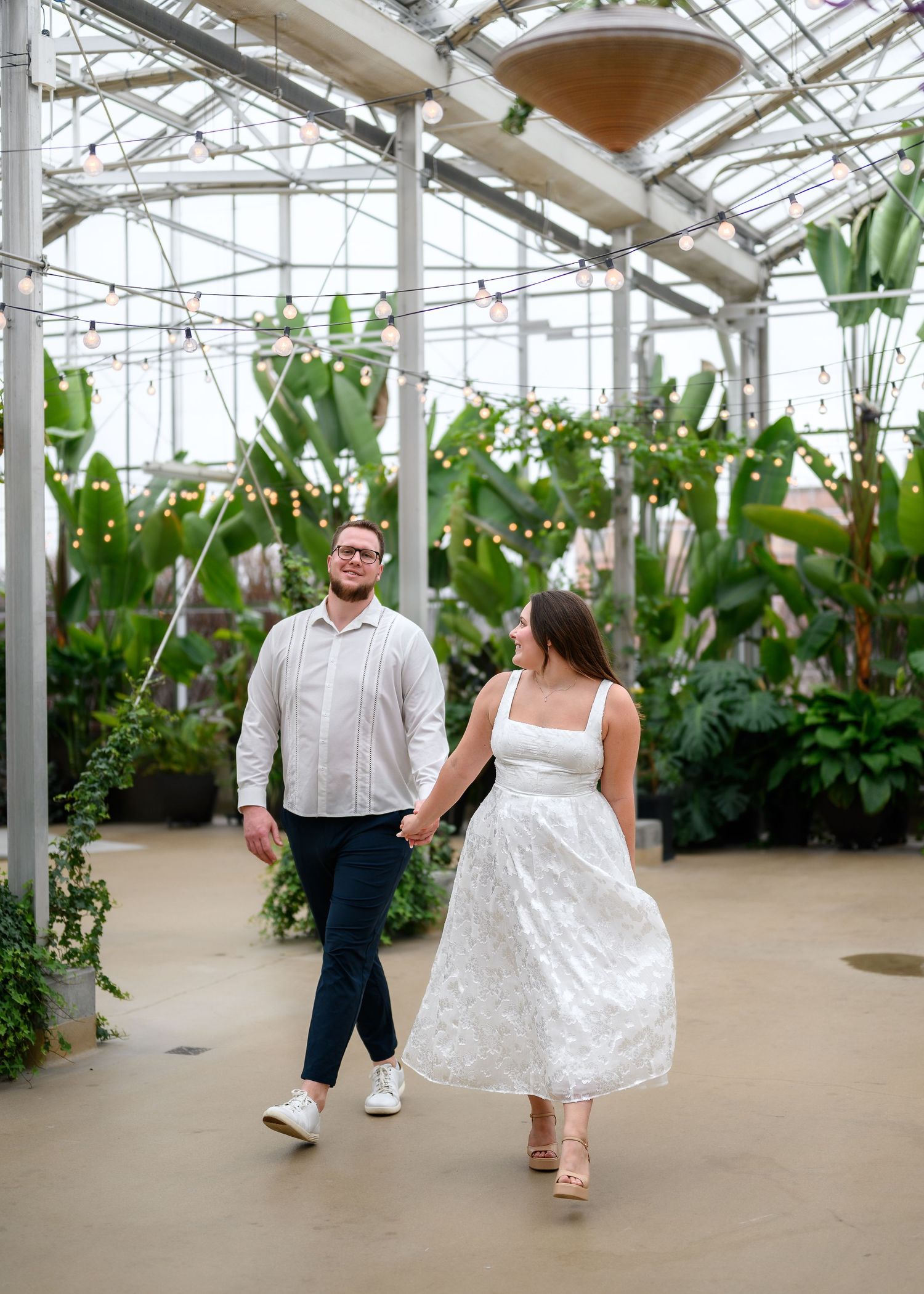 A couple dances together in a greenhouse filled with lush tropical plants and string lights overhead.