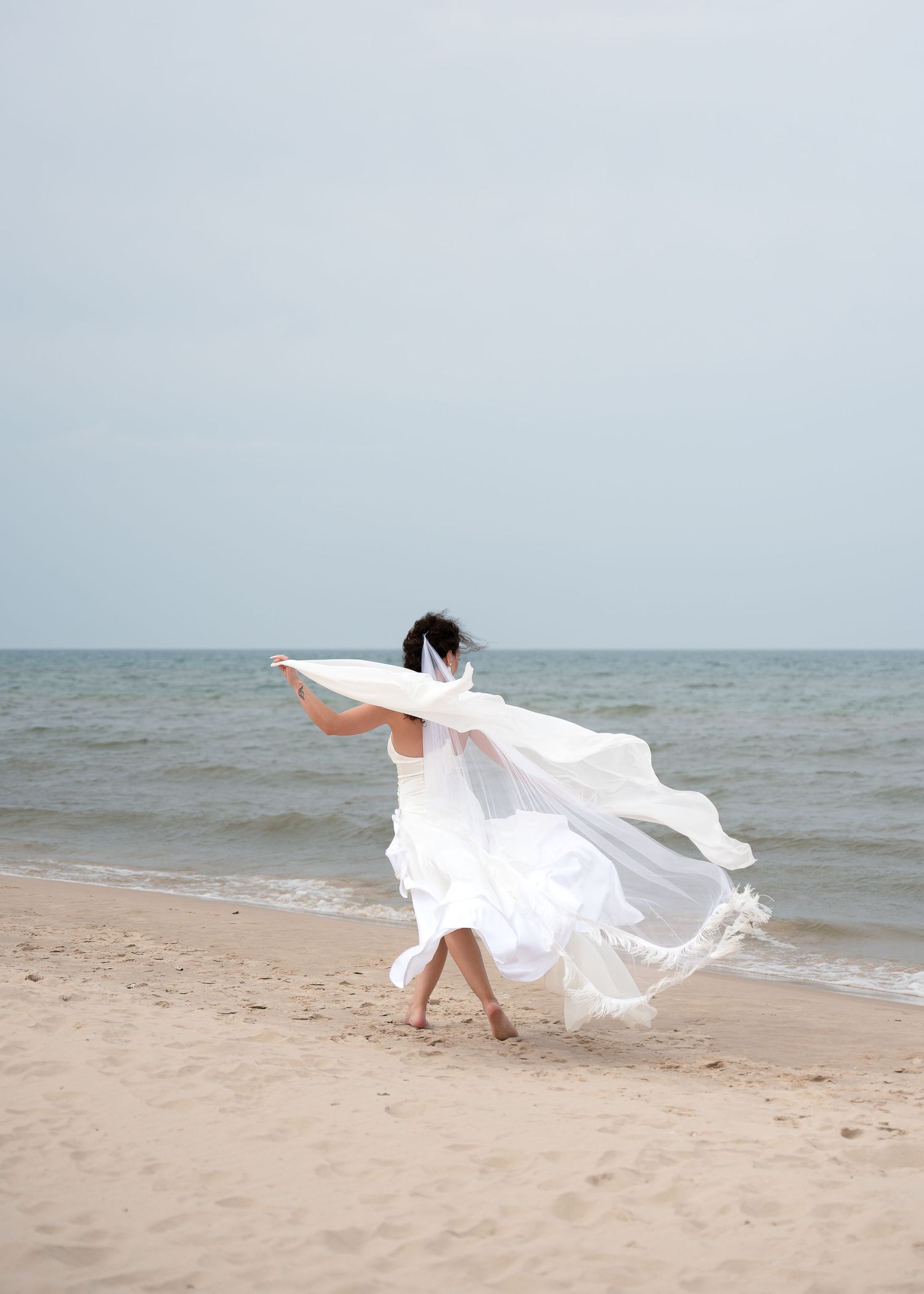 A bride in a flowing white wedding dress and veil walks barefoot on a sandy beach with the ocean in the background.