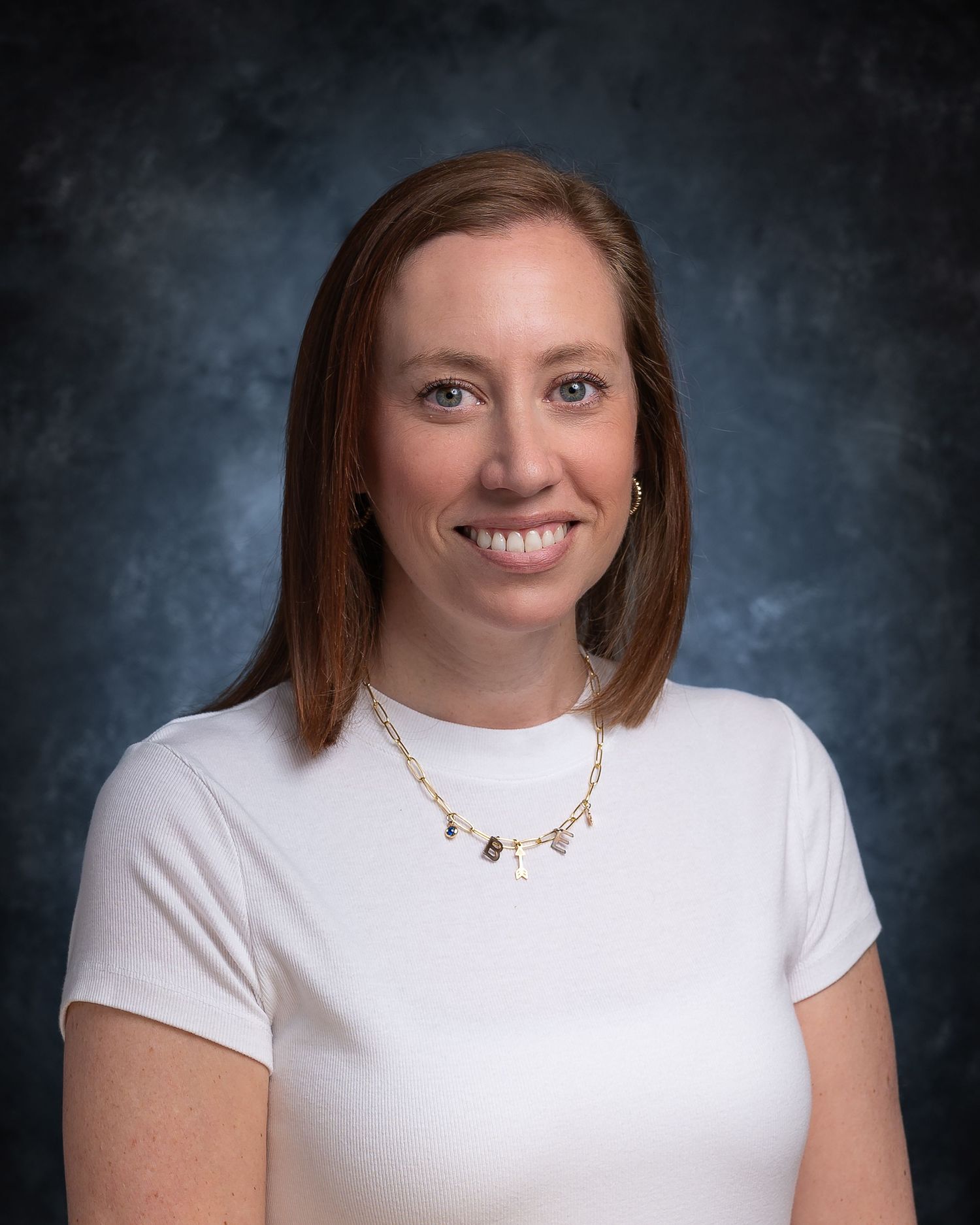 Professional headshot of a smiling woman with brown hair wearing a white top and gold necklace against a blue backdrop.