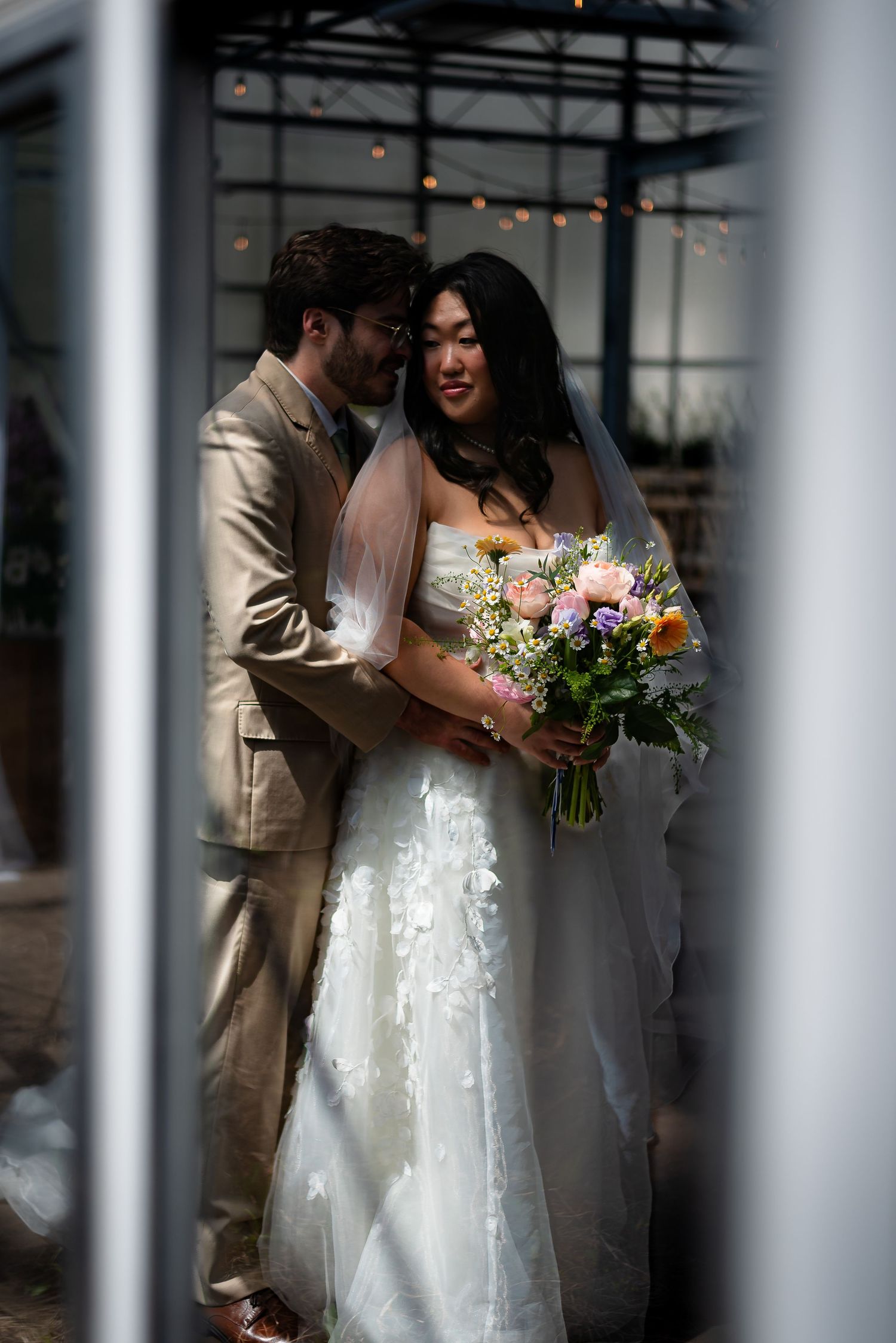 Bride in white gown holding pastel bouquet embraced by groom in tan suit, romantic wedding portrait.