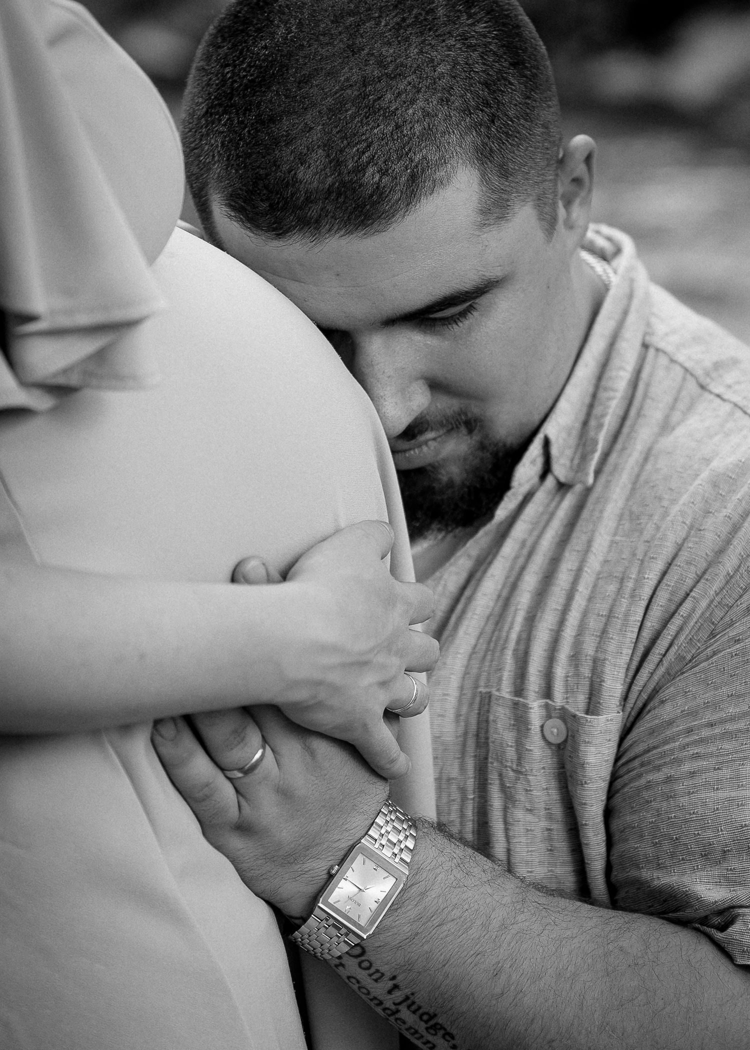 Black and white photo of a man tenderly resting his head on a pregnant woman's belly, holding her hands.