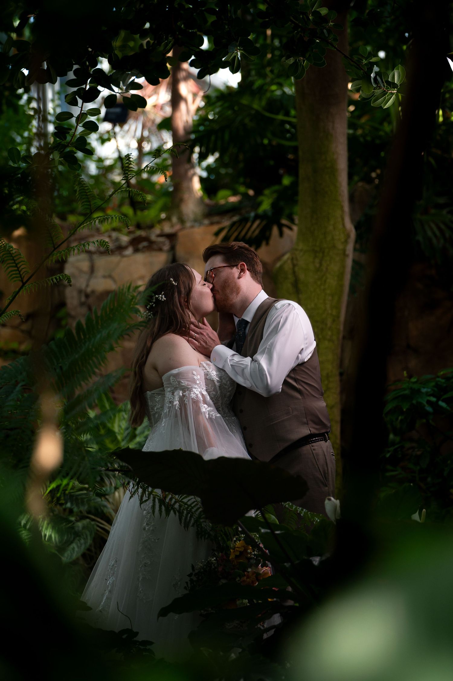 A bride and groom share a romantic kiss surrounded by lush green ferns and trees in an enchanting garden setting.