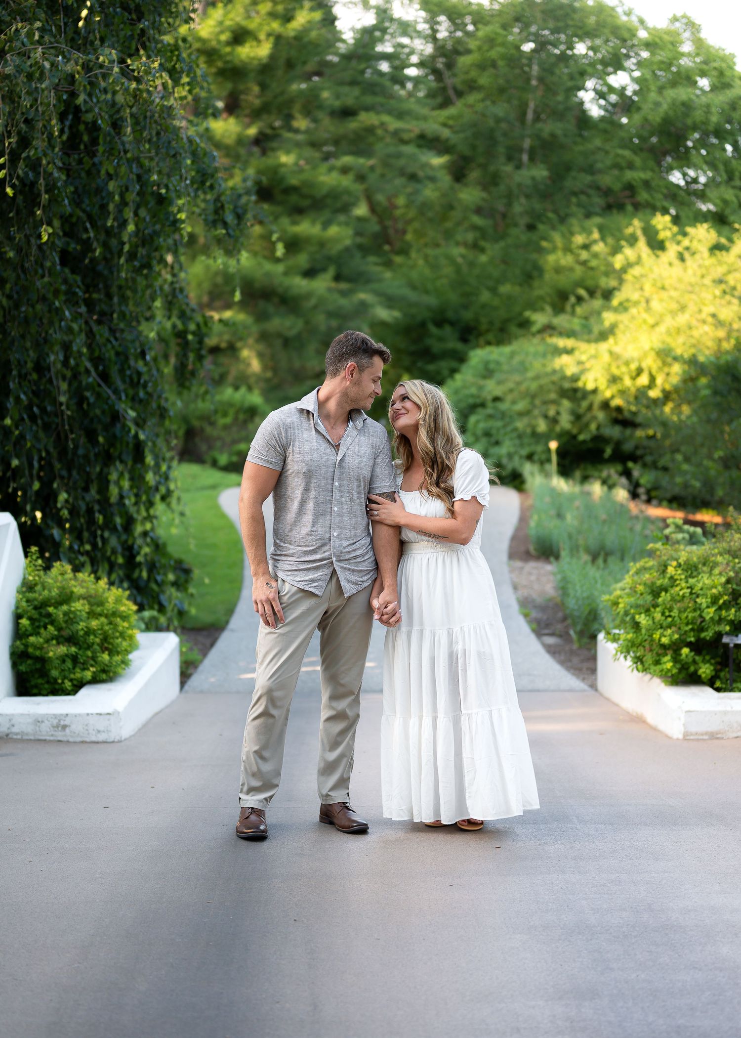 Couple in love gazes at each other on a garden path, man in gray shirt and woman in white maxi dress, lush greenery background.