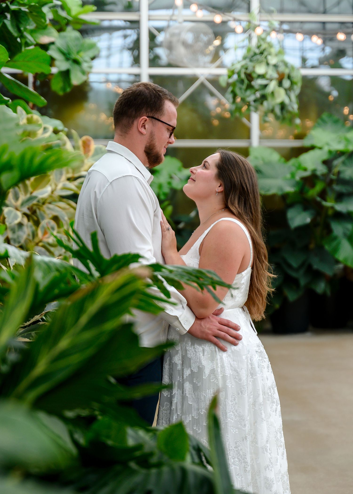 Romantic couple embraces in a lush greenhouse setting surrounded by green plants and natural light.