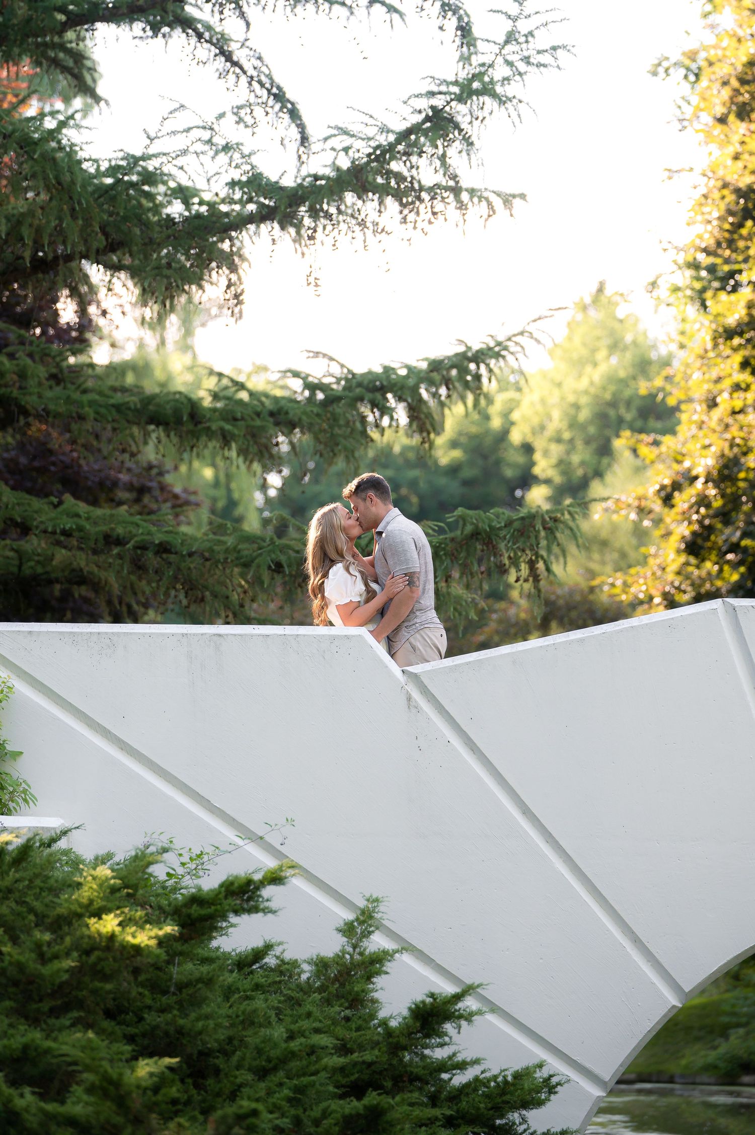 Couple sharing a romantic kiss on a white architectural structure surrounded by lush green trees in a garden setting.