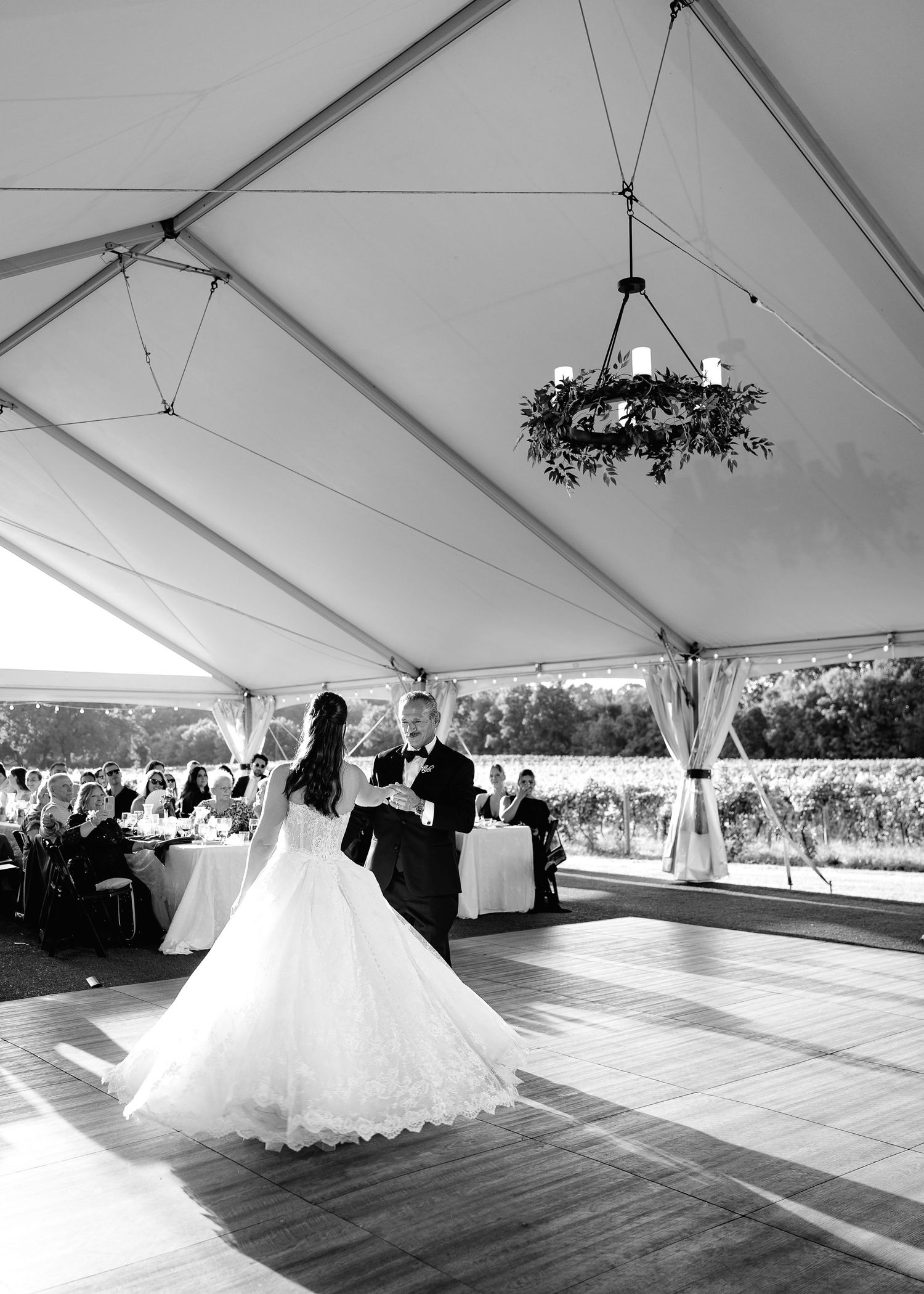 Black and white photo of bride and groom sharing first dance under elegant tent with floral chandelier.