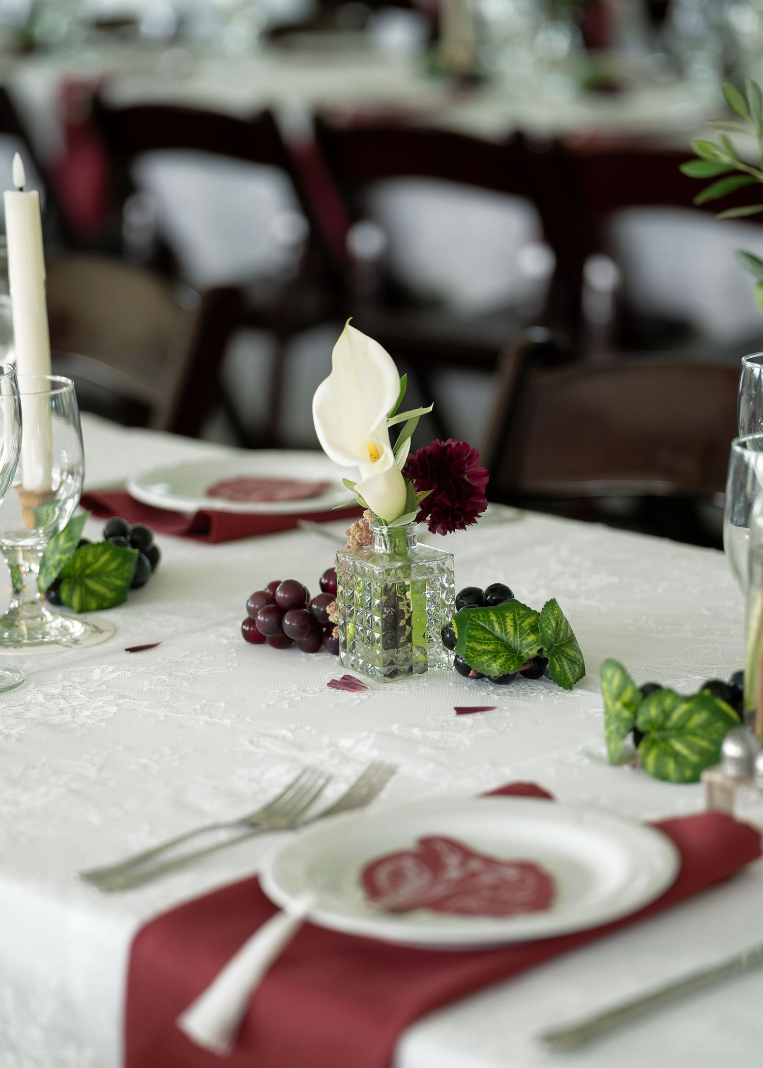 Elegant wedding reception table with white calla lily centerpiece, burgundy napkins, candles, and grape accents on white linen.