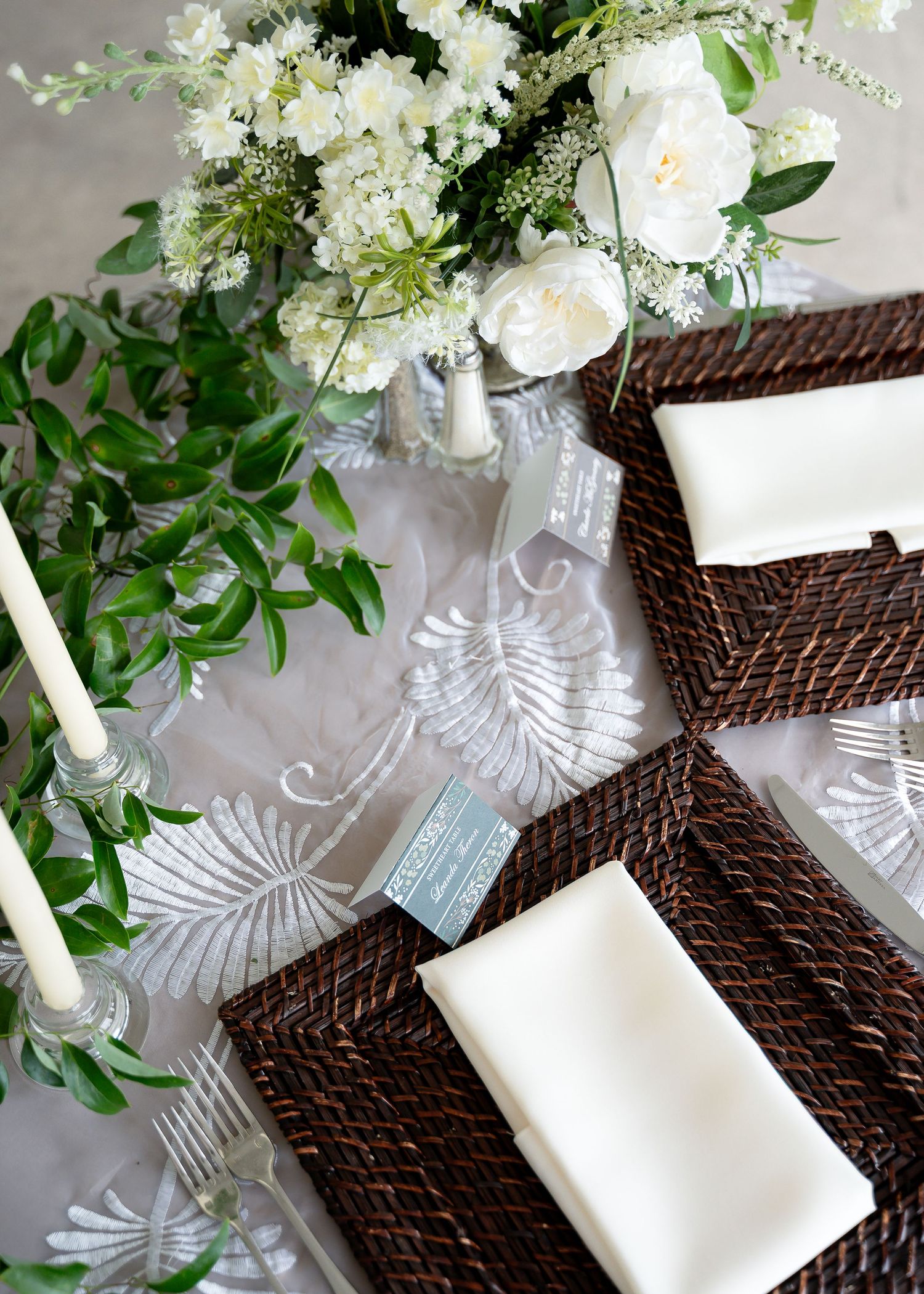 Elegant wedding table setting with woven chargers, white linen napkins, white floral centerpiece, and greenery on embroidered tablecloth.