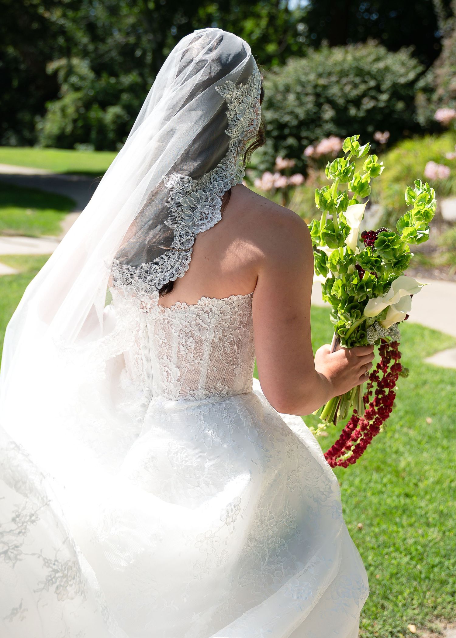 Bride in lace wedding dress and veil holding bouquet of white flowers and red beads in sunny garden.