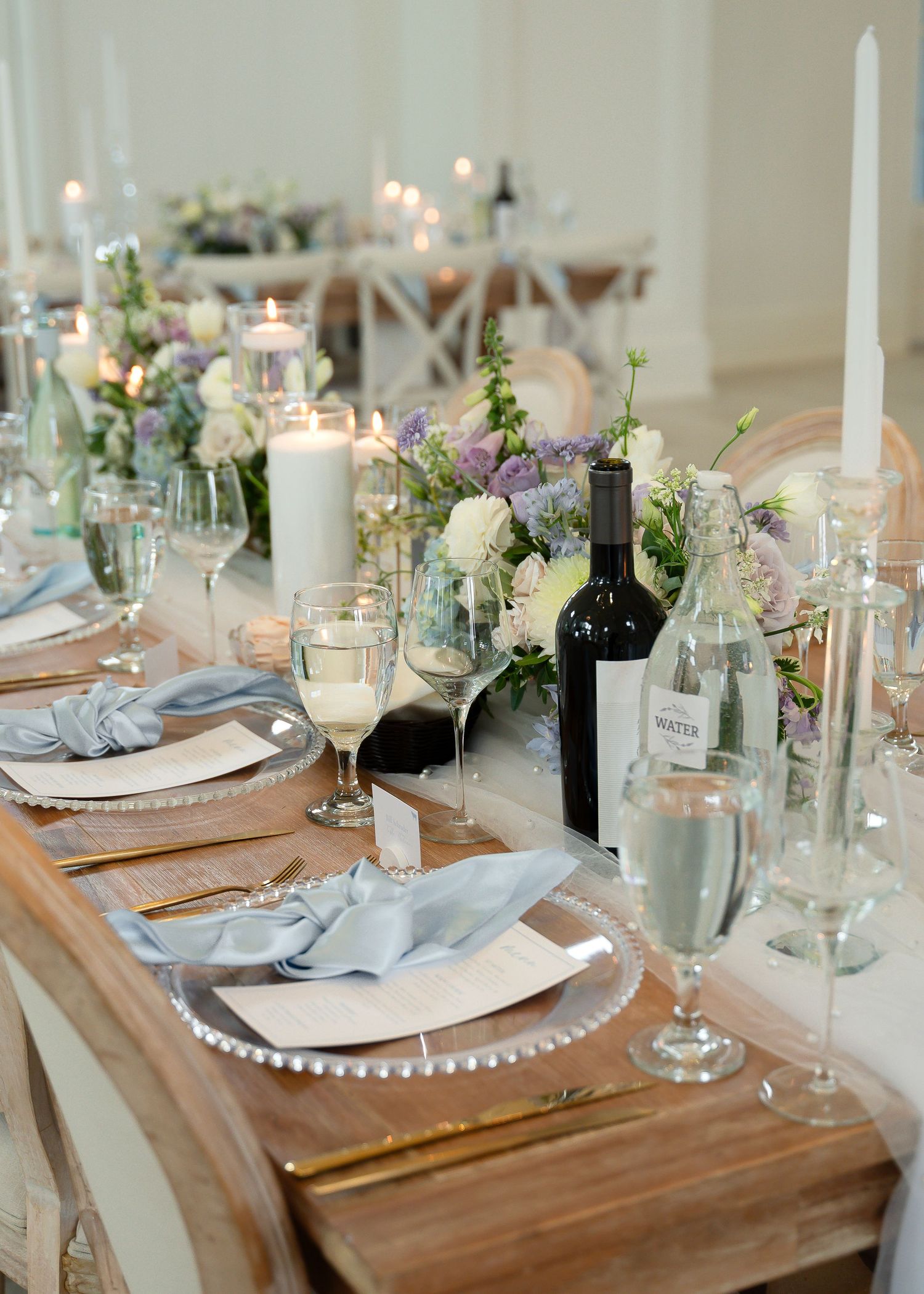 Elegant wedding table setting with dusty blue napkins, floral centerpieces, candles, and wine glasses on a rustic wood table.