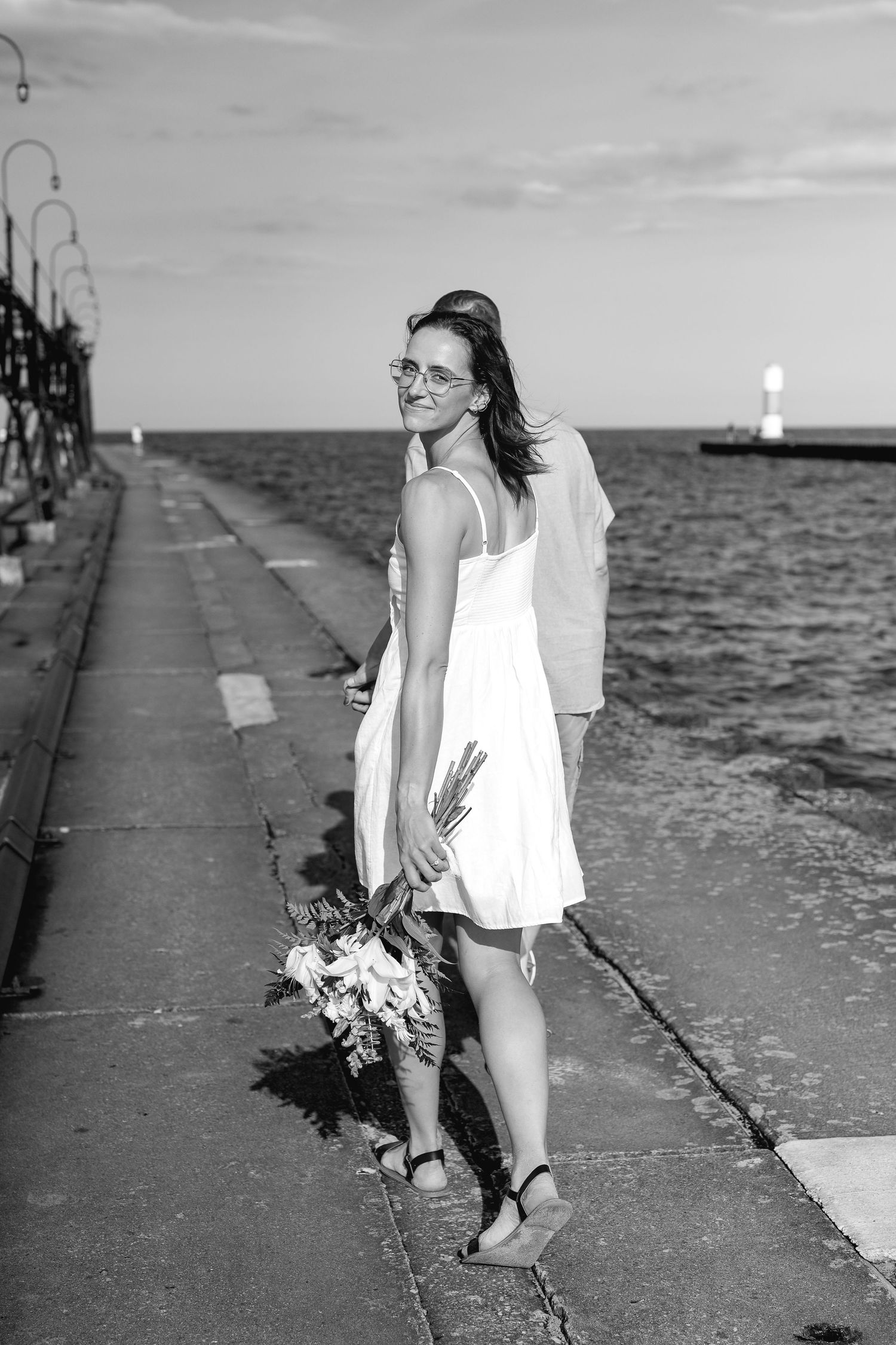 Woman in white dress holding bouquet on waterfront pier, black and white photo.